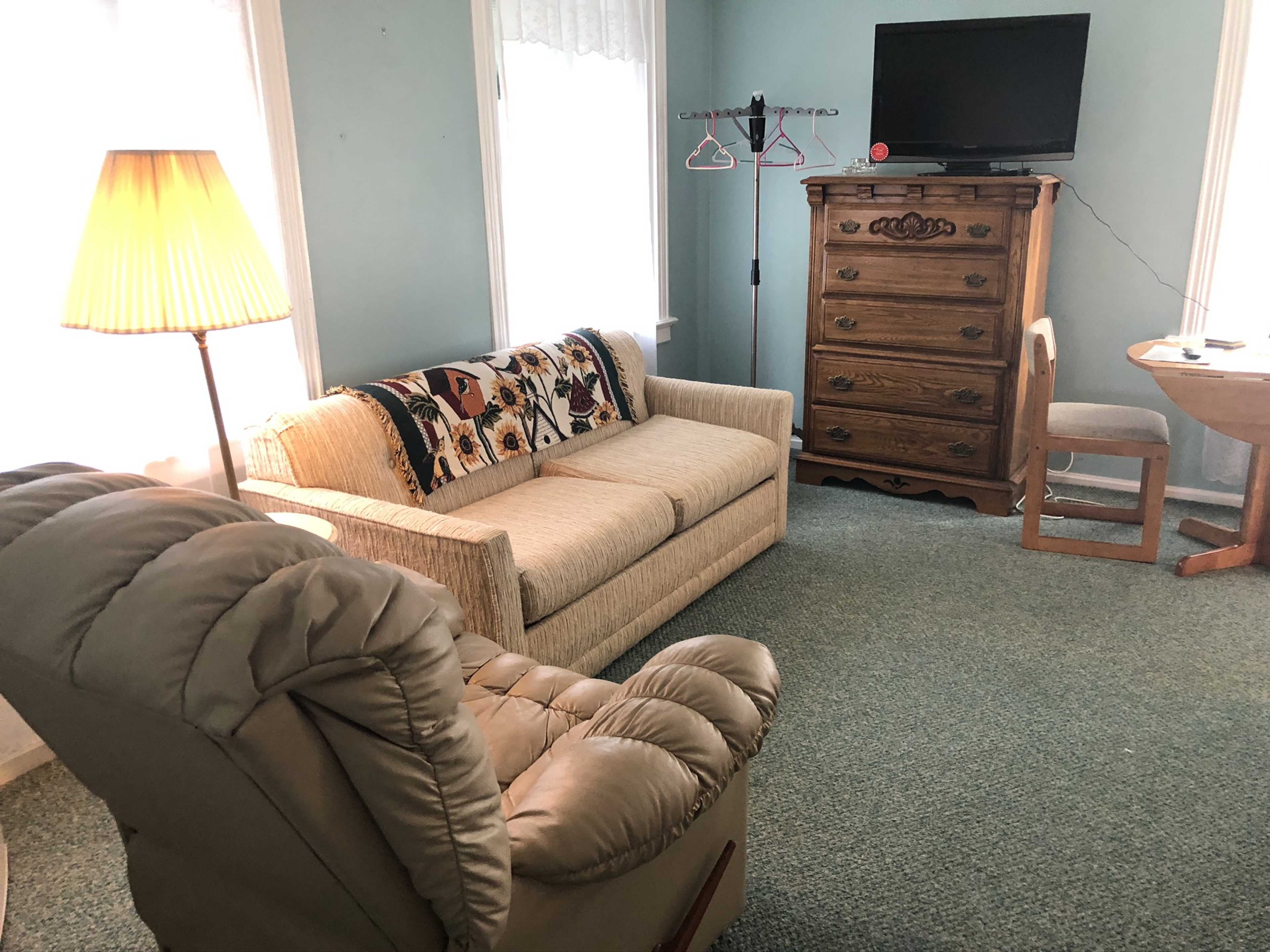 A cozy room with a beige sofa, a recliner, a wooden dresser, a small table, and a television mounted on the wall.