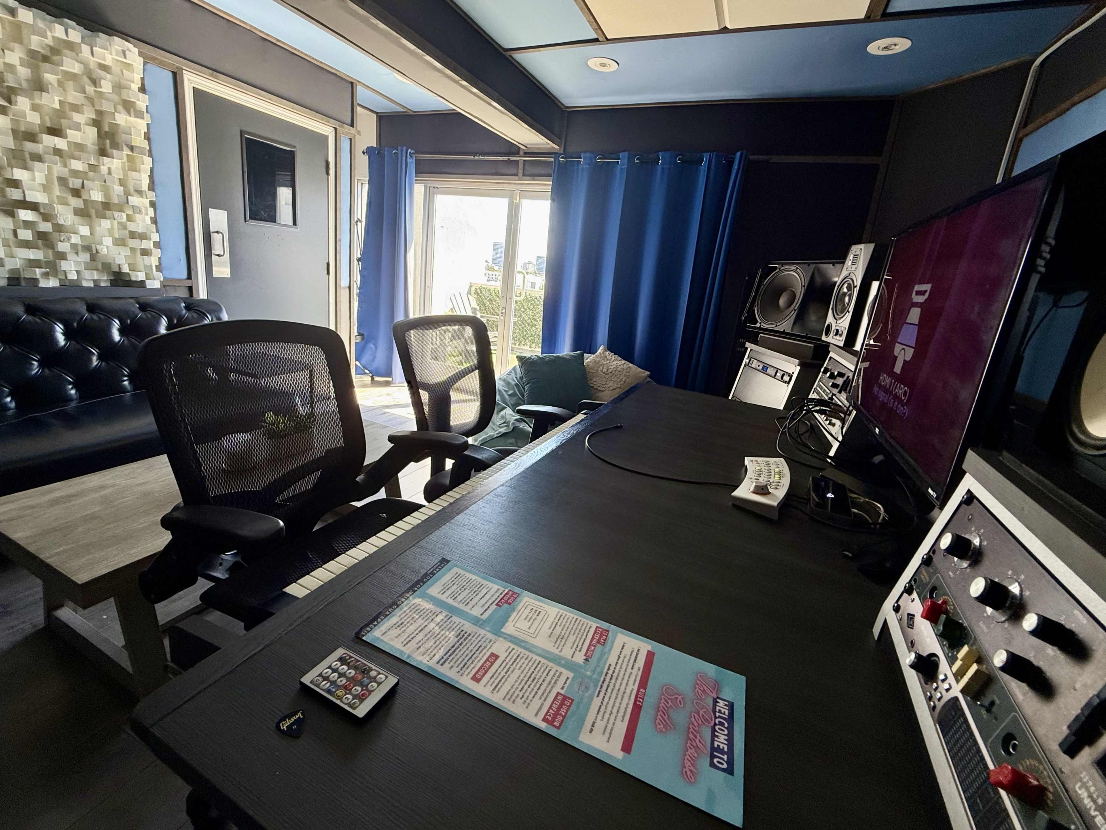 A modern music studio is set up with a desk featuring a keyboard and monitor, complemented by a comfortable seating area and blue curtains.