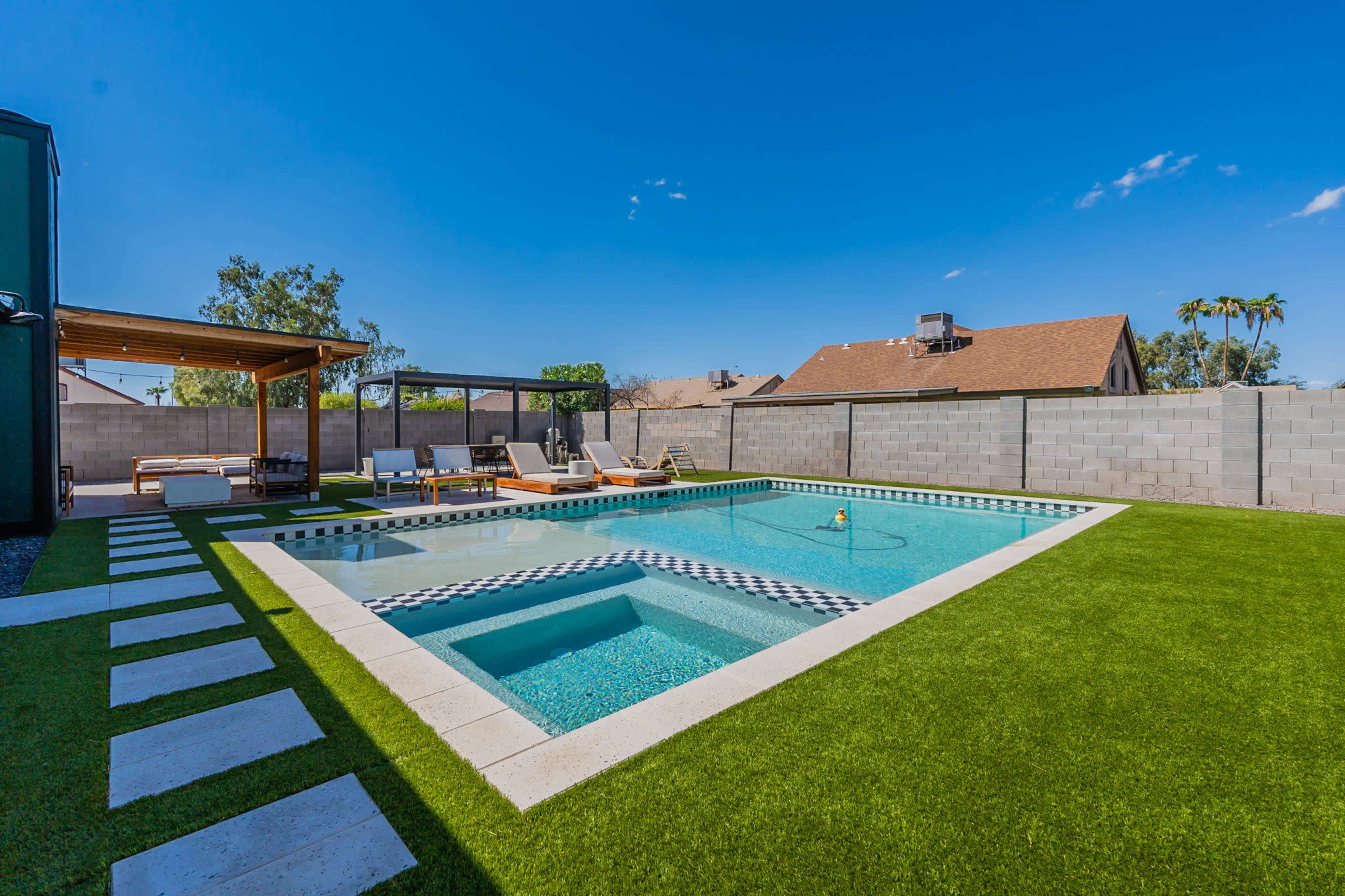 The image showcases a rectangular pool surrounded by artificial grass and stone pathways, with lounge chairs positioned nearby.