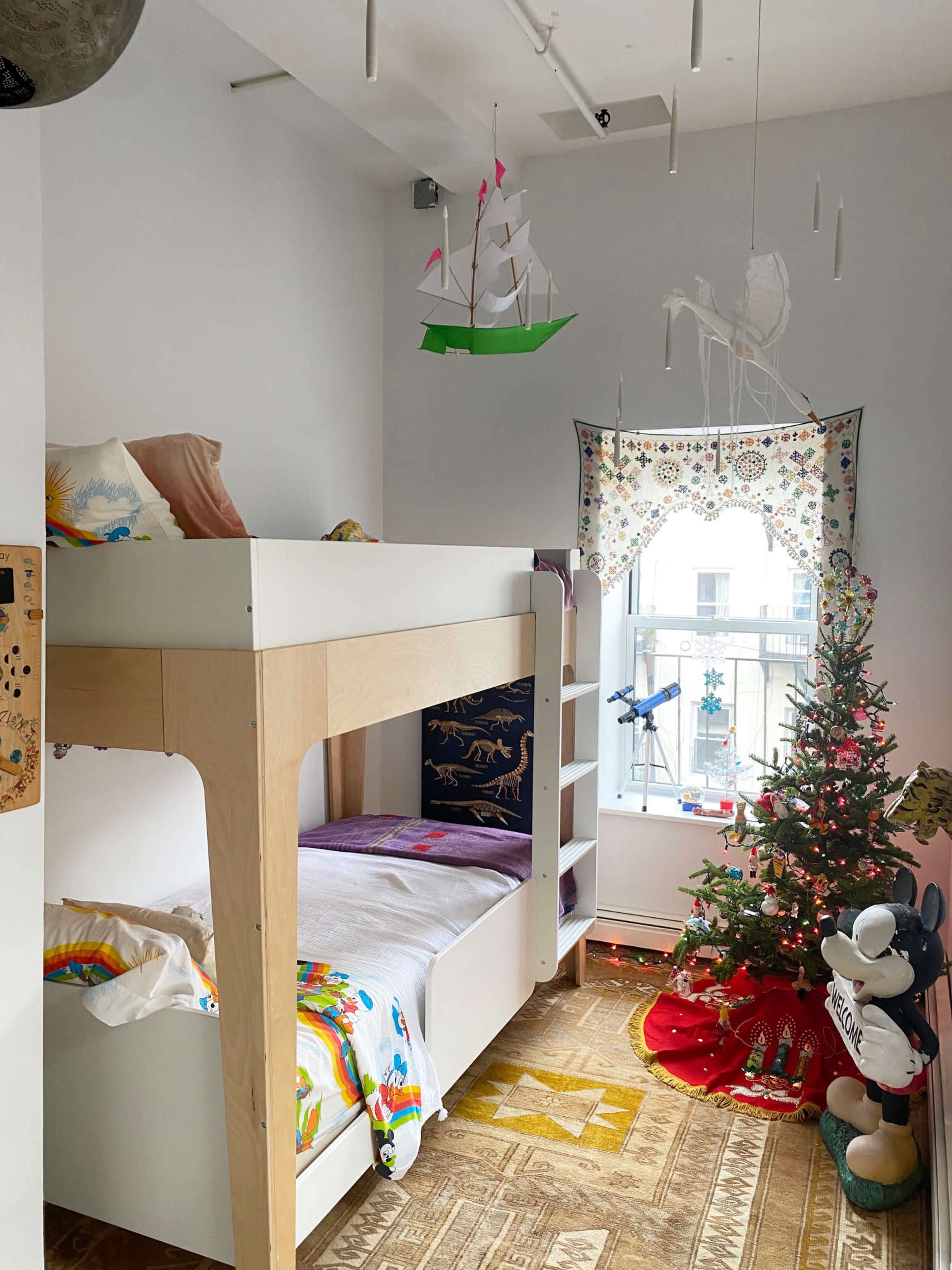 A children's bedroom featuring a lofted bed with a lower sleeping area, a decorated Christmas tree in the corner, and a window adorned with ornamental curtains.