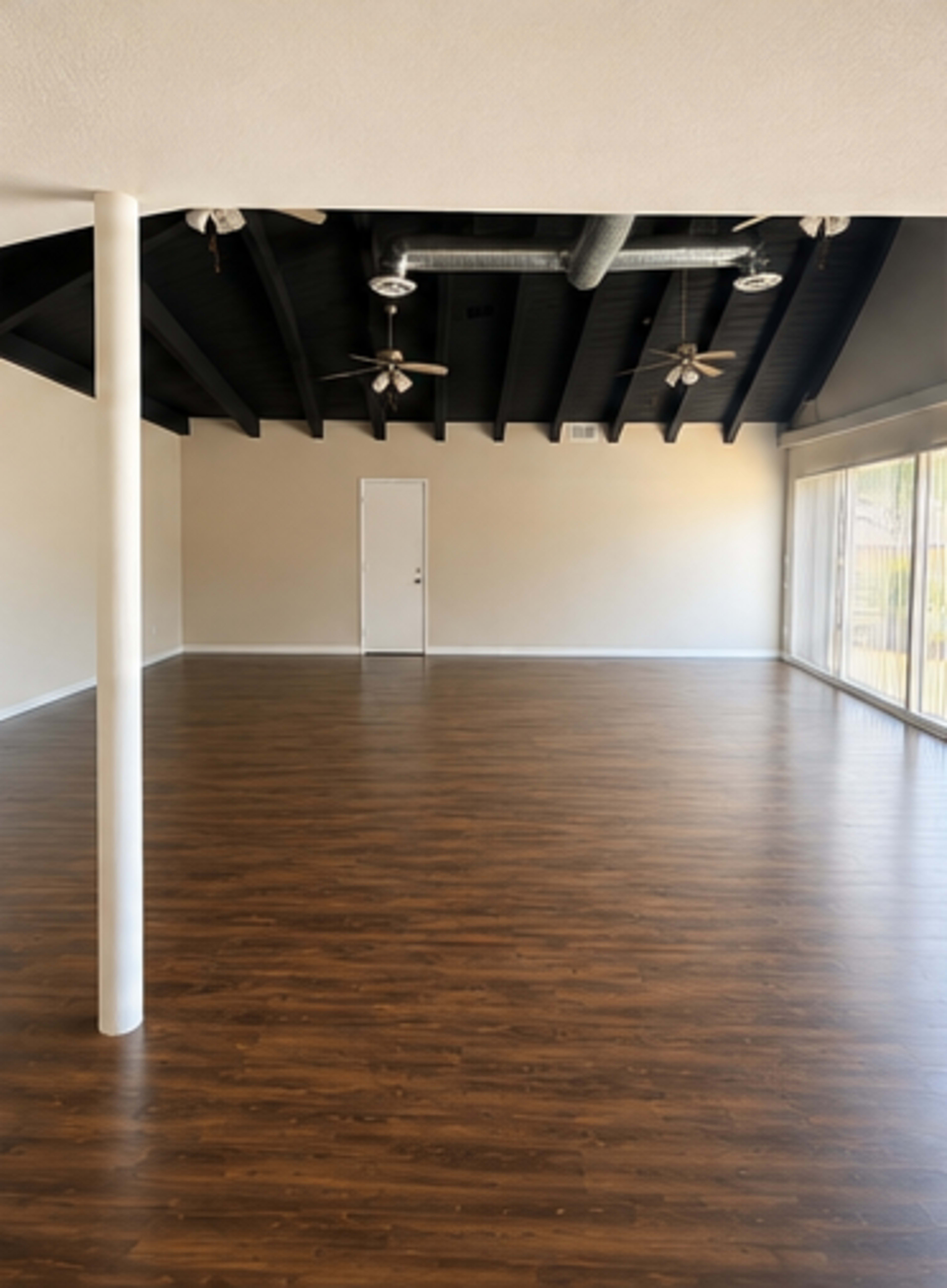 The image shows an empty room with polished wooden floors, exposed beam ceilings, and large windows along one wall.