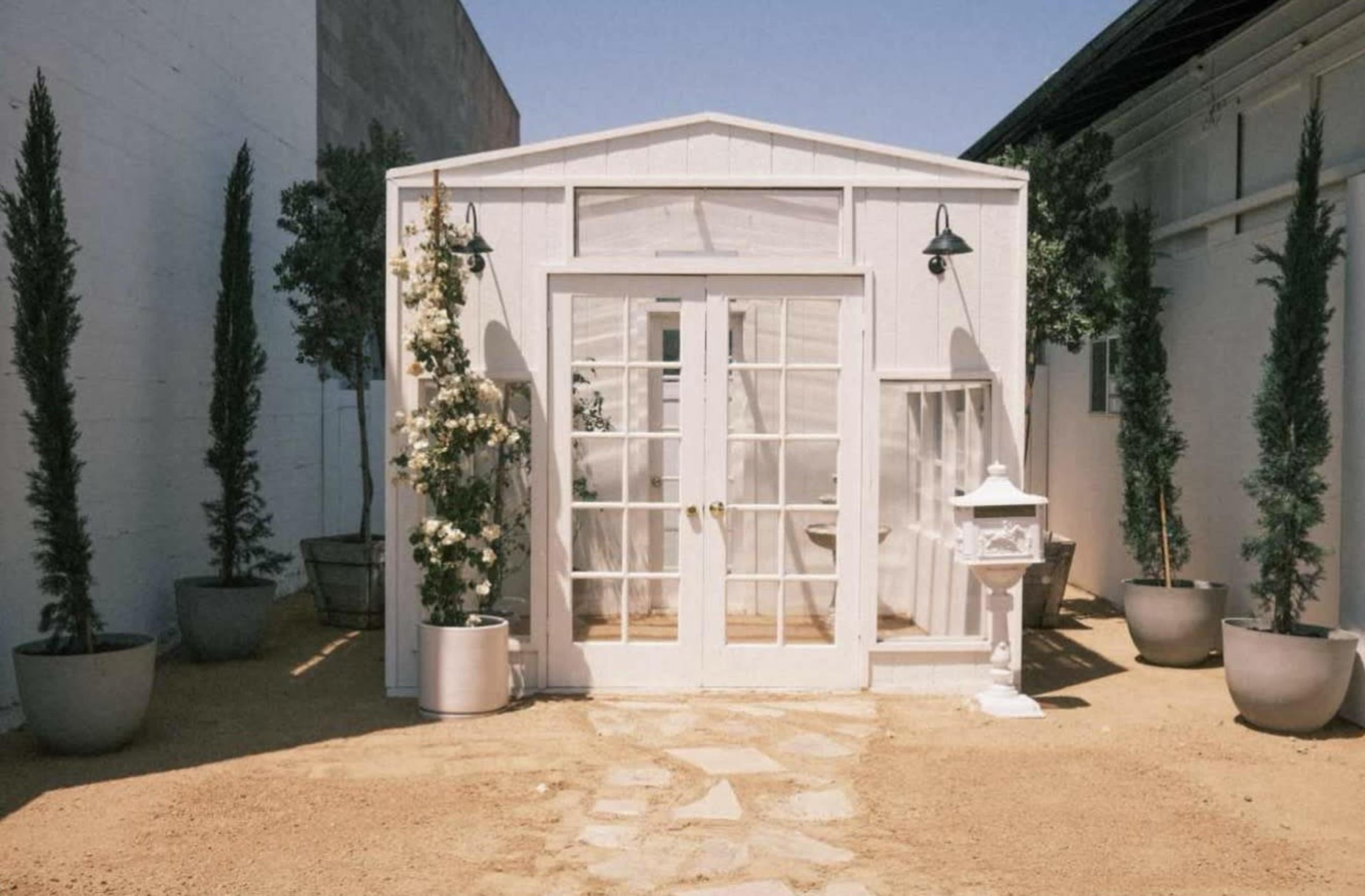 A small white structure with large windows and a double door stands on a sandy path, surrounded by potted plants and tall trees.