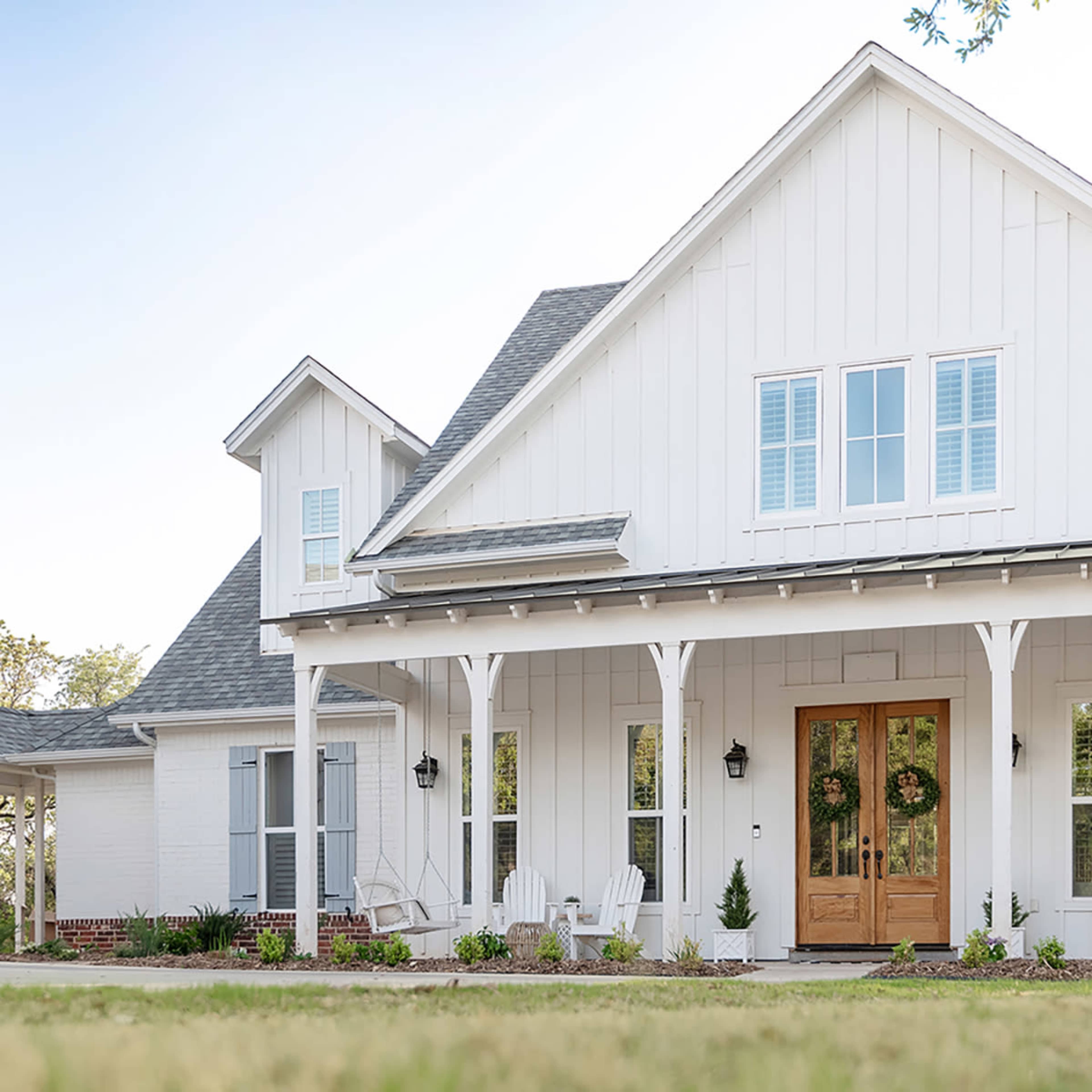 A modern white farmhouse features a large front porch with wooden doors and rocking chairs on either side.