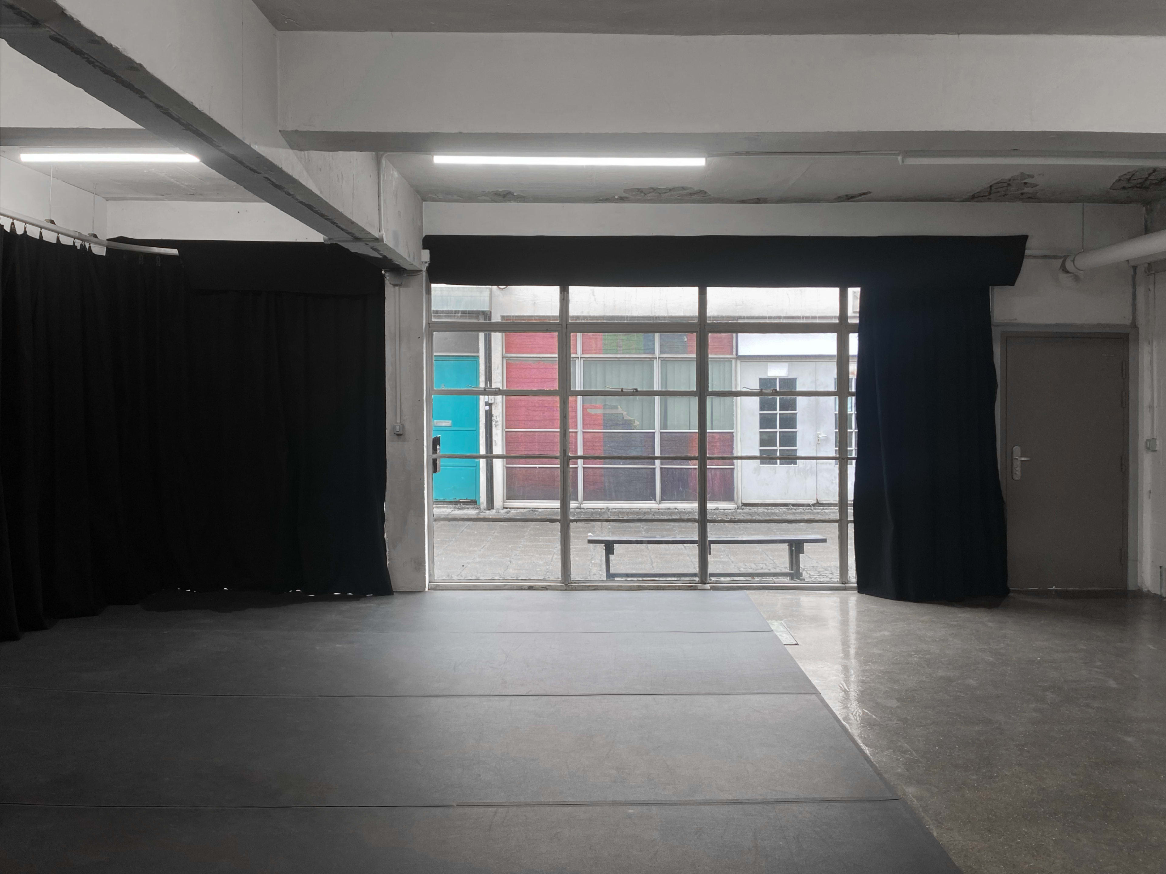 A spacious room with a dark floor, black curtains, and large windows revealing an outdoor view with colorful buildings.