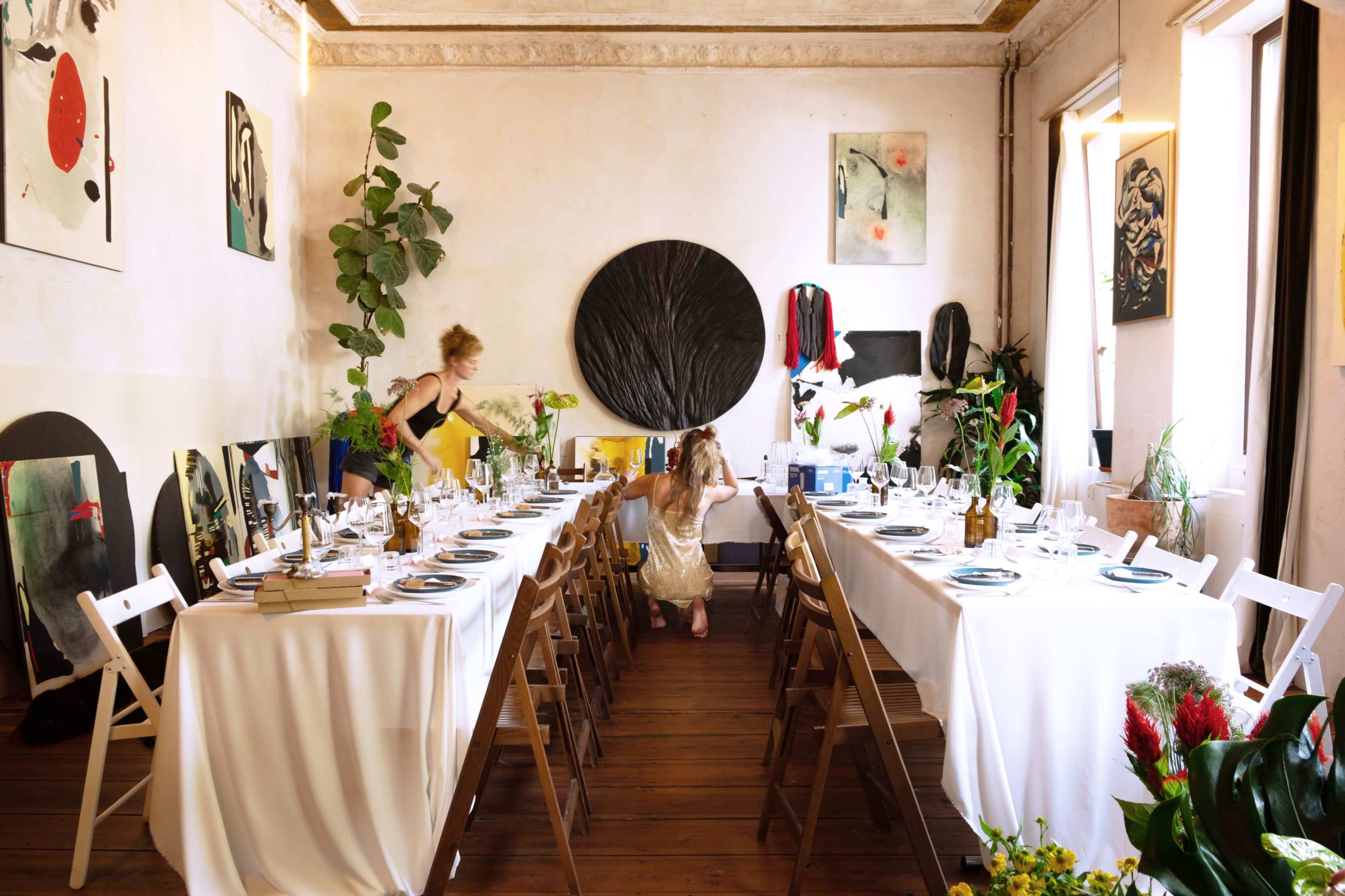 A dining area features a long table set with white tablecloths, surrounded by wooden chairs, and decorated with various plants and artworks.