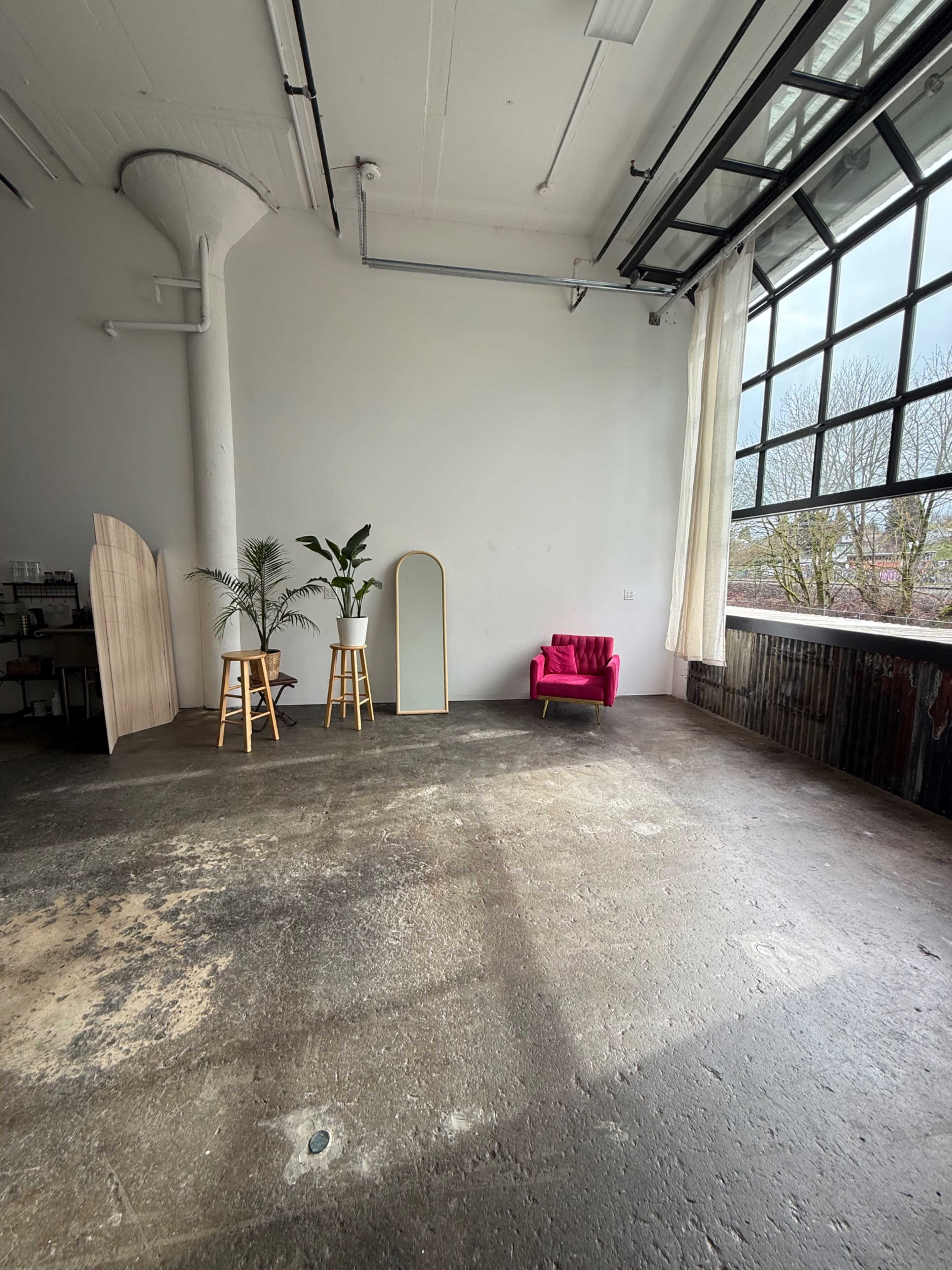 The image shows a spacious interior with a concrete floor, large windows, a pink chair, two wooden stools, a floor mirror, and two potted plants.