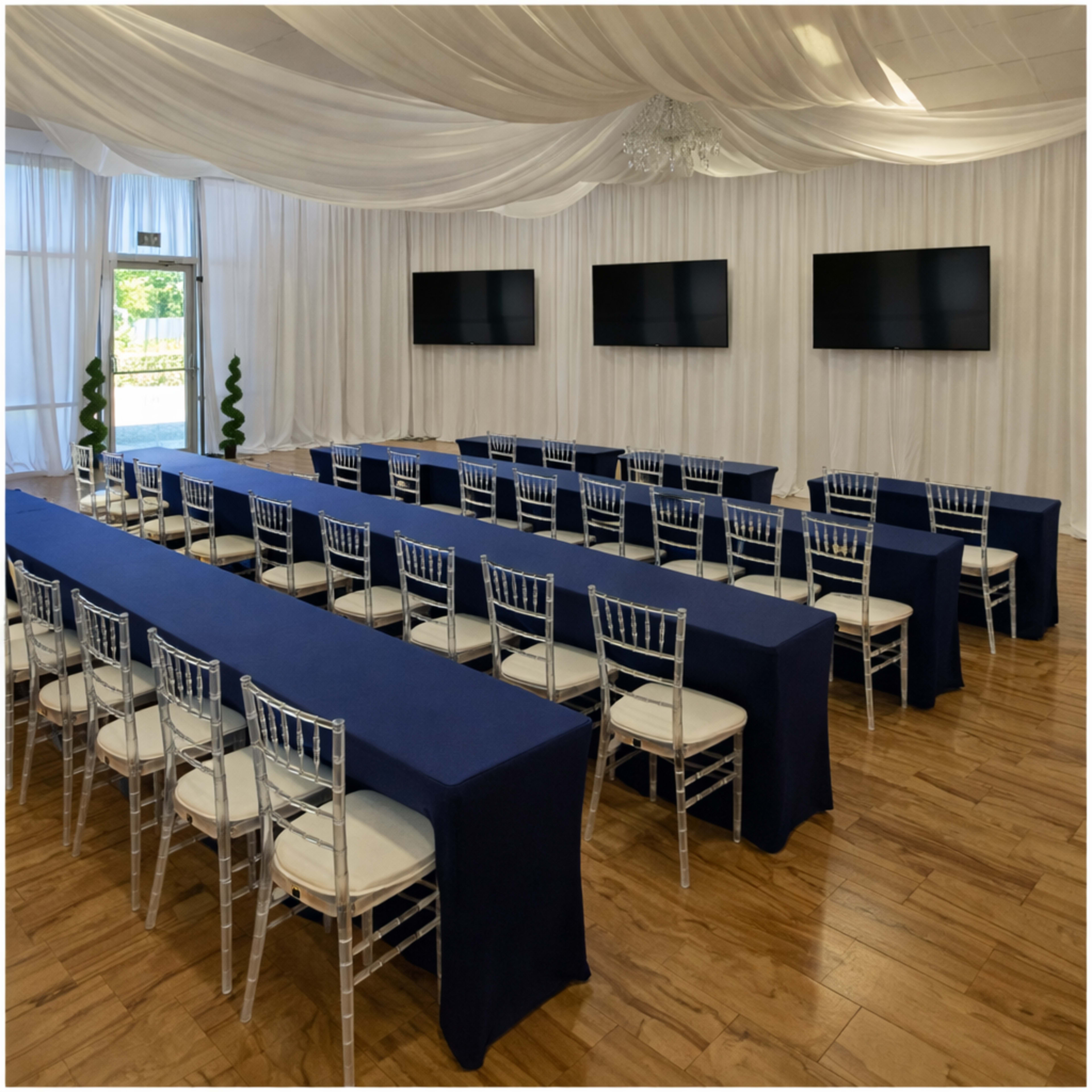 The image shows a spacious event room set up with long tables covered in blue cloth, arranged in rows, with clear chairs and three mounted screens on the wall.