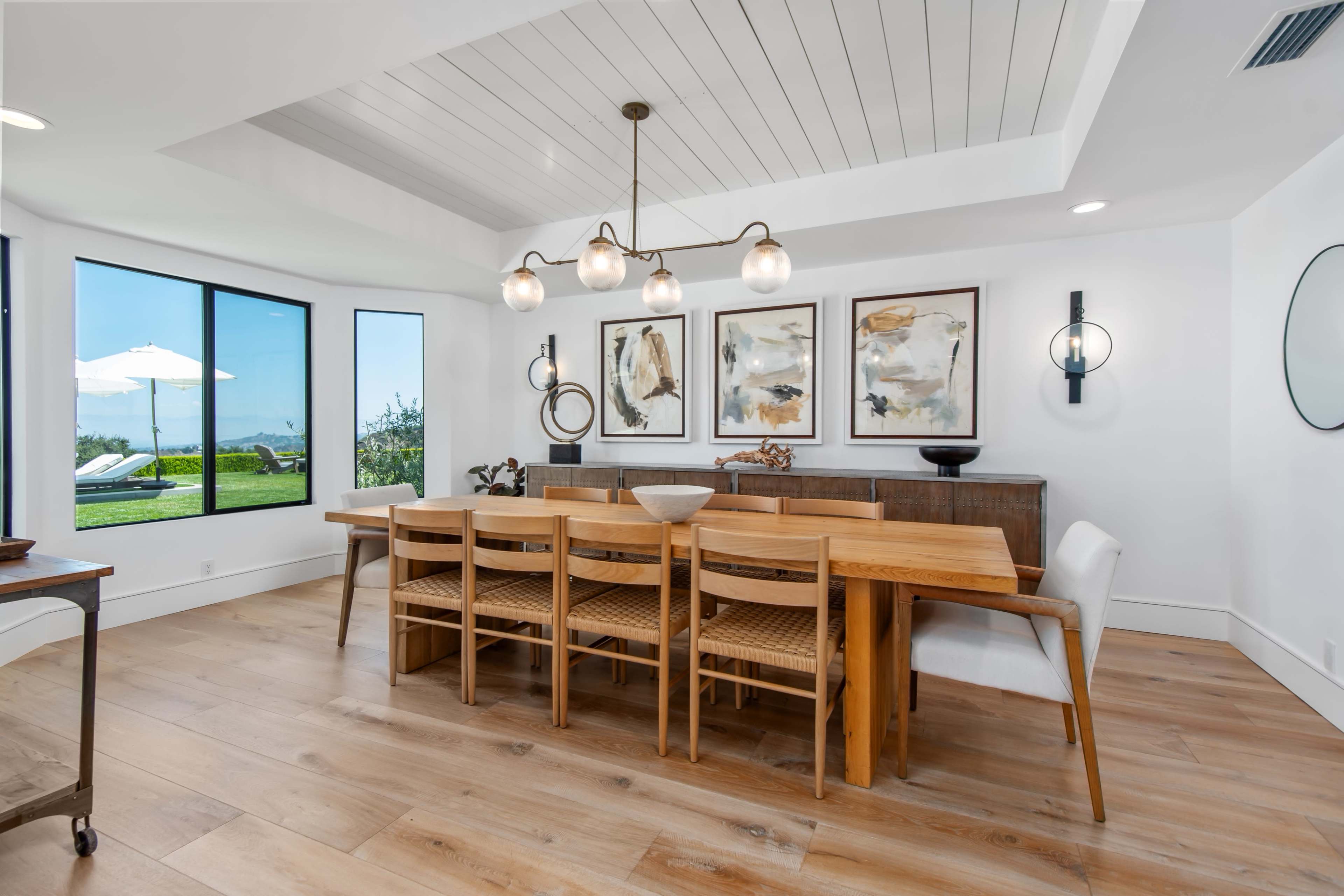 A spacious dining area features a long wooden table surrounded by chairs, with large windows offering views of a landscaped outdoor space.