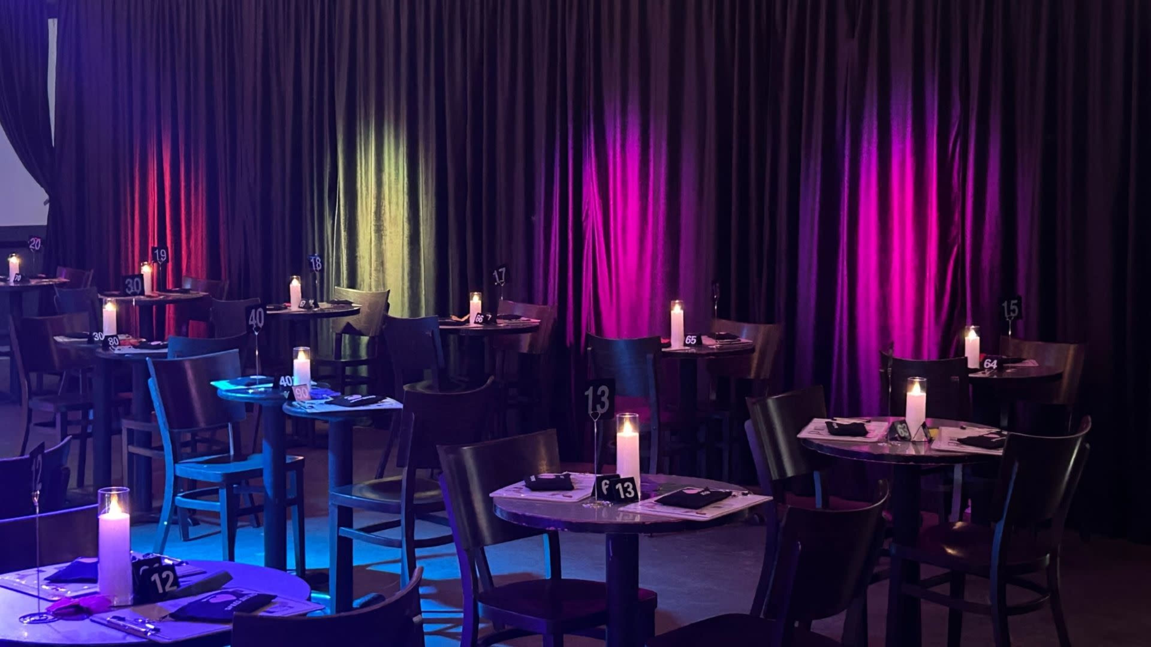 The image shows a dimly lit dining area with multiple tables, each set for guests and adorned with candles, against a backdrop of colorful illuminated curtains.