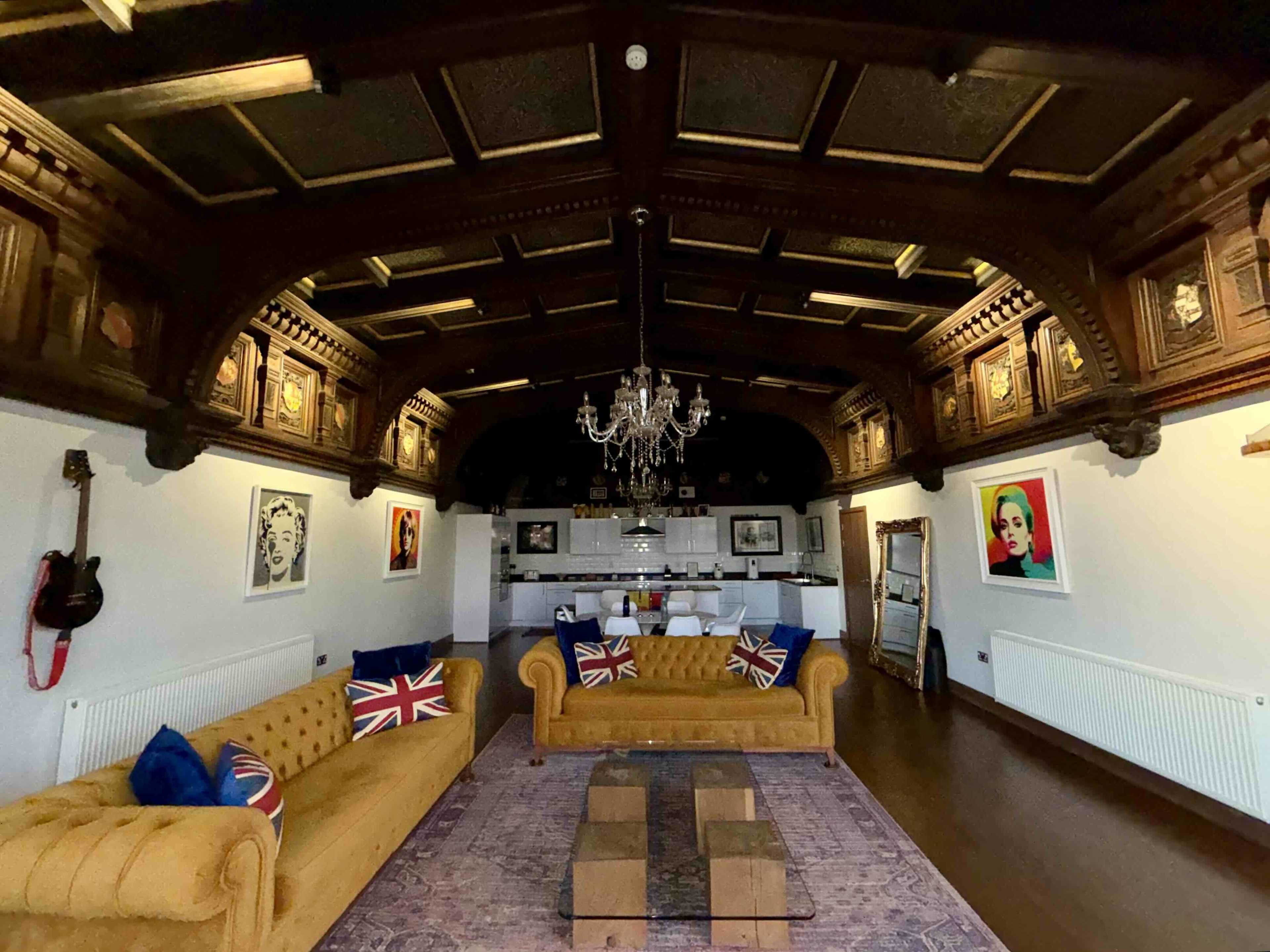 The image shows a spacious living area with a high, ornate wooden ceiling, two yellow sofas adorned with Union Jack pillows, and a view of a kitchen in the background.