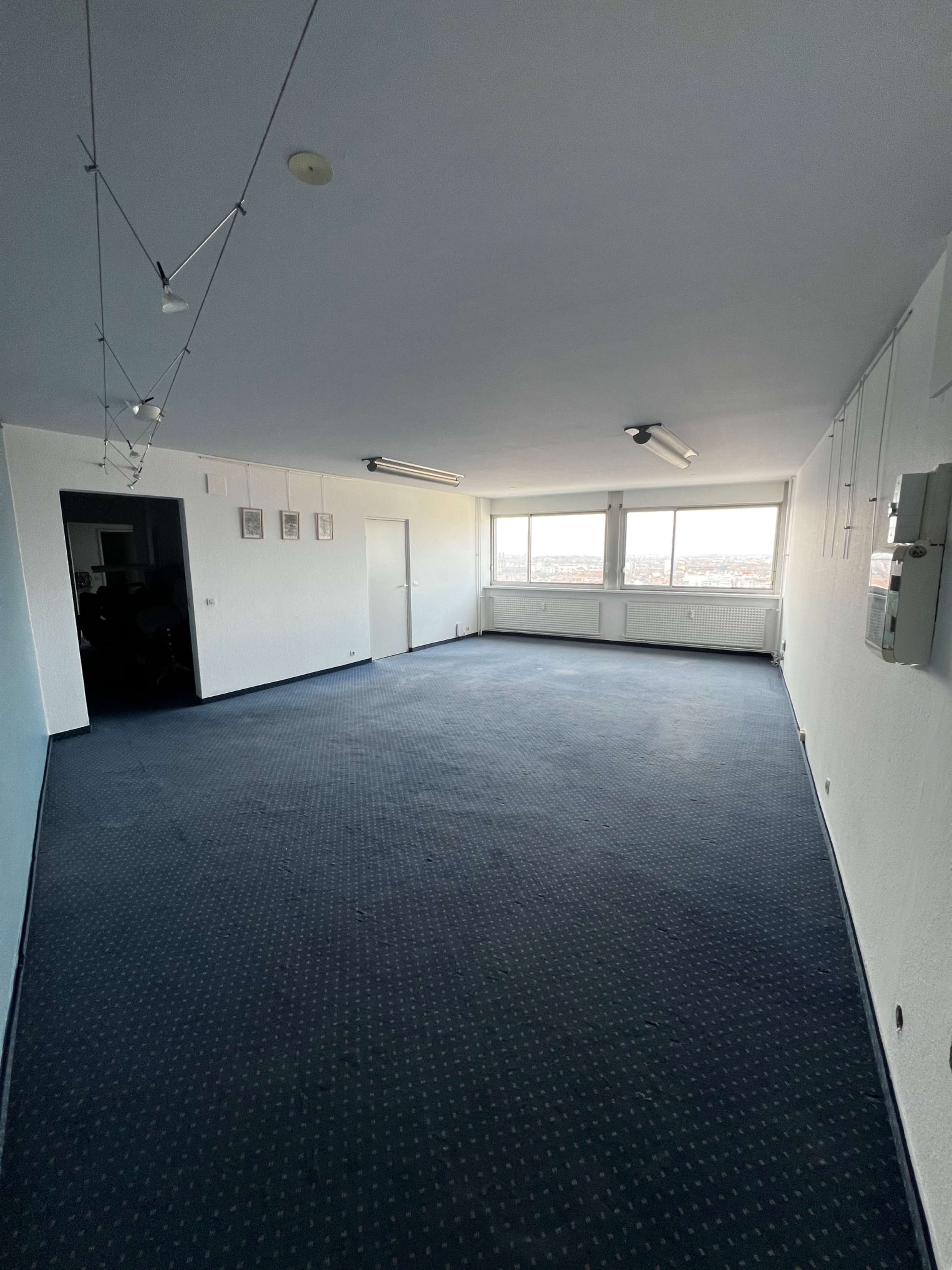 The image shows a sparsely furnished office space with blue carpeting and large windows that provide natural light.