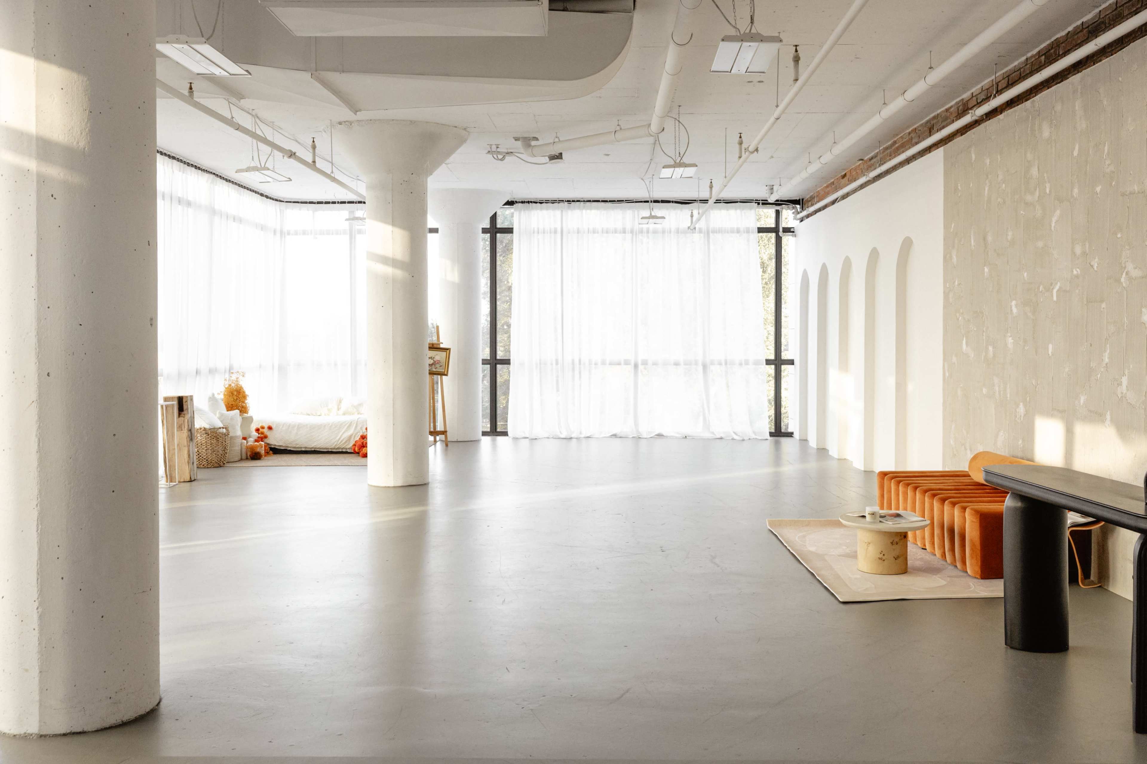 The image shows a spacious, minimalist interior featuring large windows with sheer curtains, a concrete floor, and a mixture of furniture pieces, including a sofa and a coffee table.