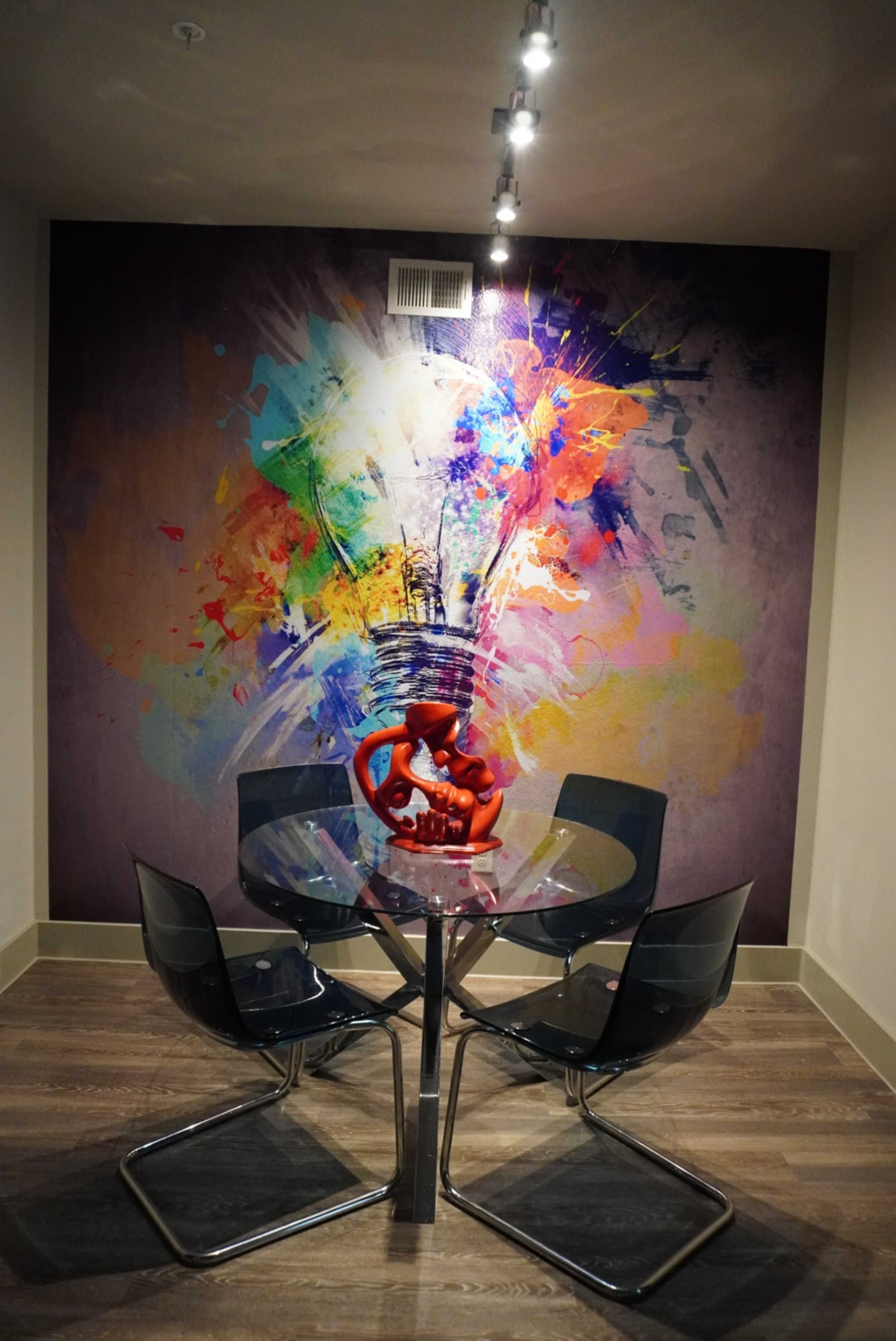 A modern dining area features a glass table surrounded by transparent chairs, with a colorful abstract mural and a red sculpture as focal points.