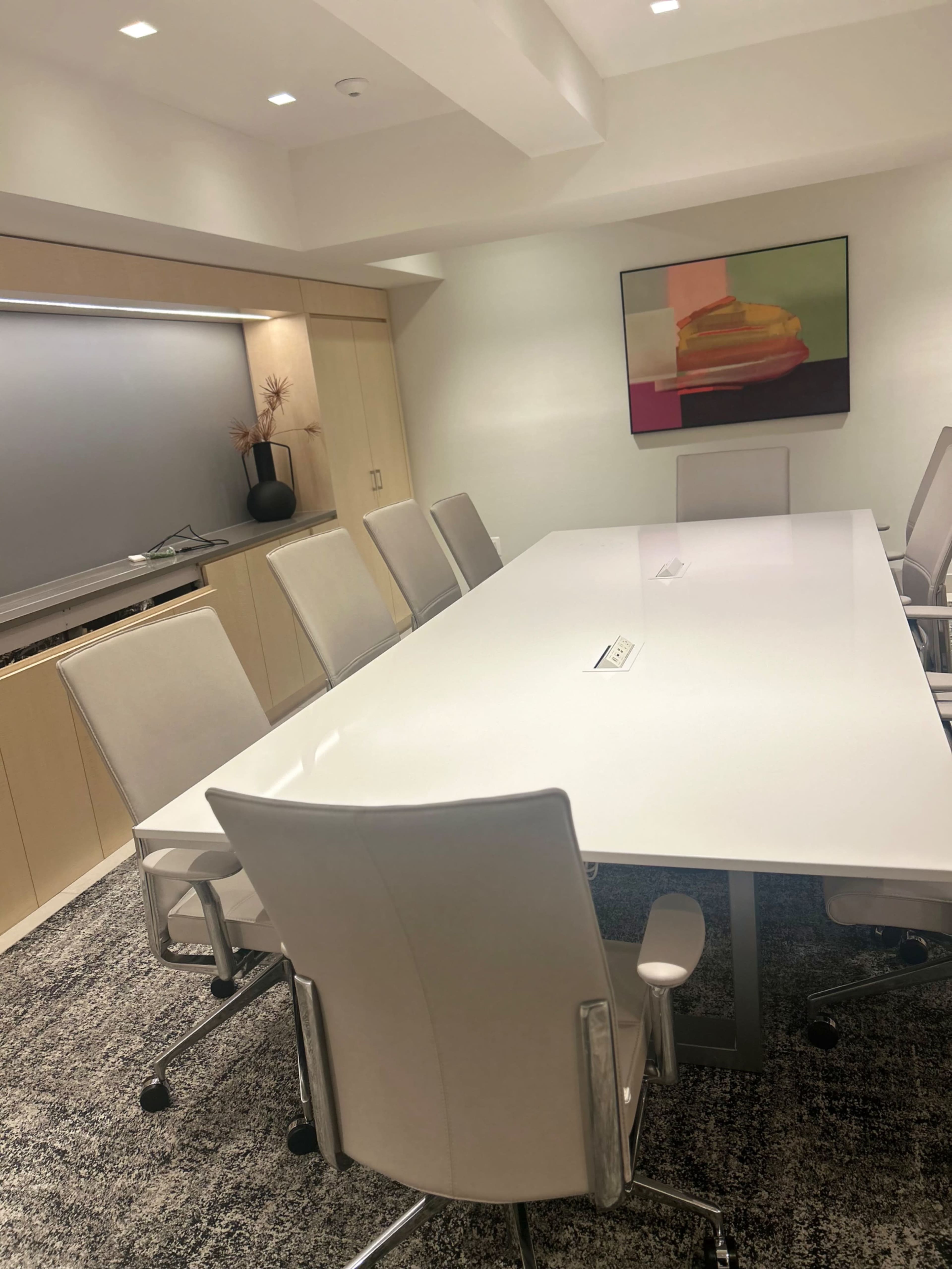 A modern conference room features a large white table surrounded by several light-colored chairs, with a wall-mounted screen and minimalist decor.