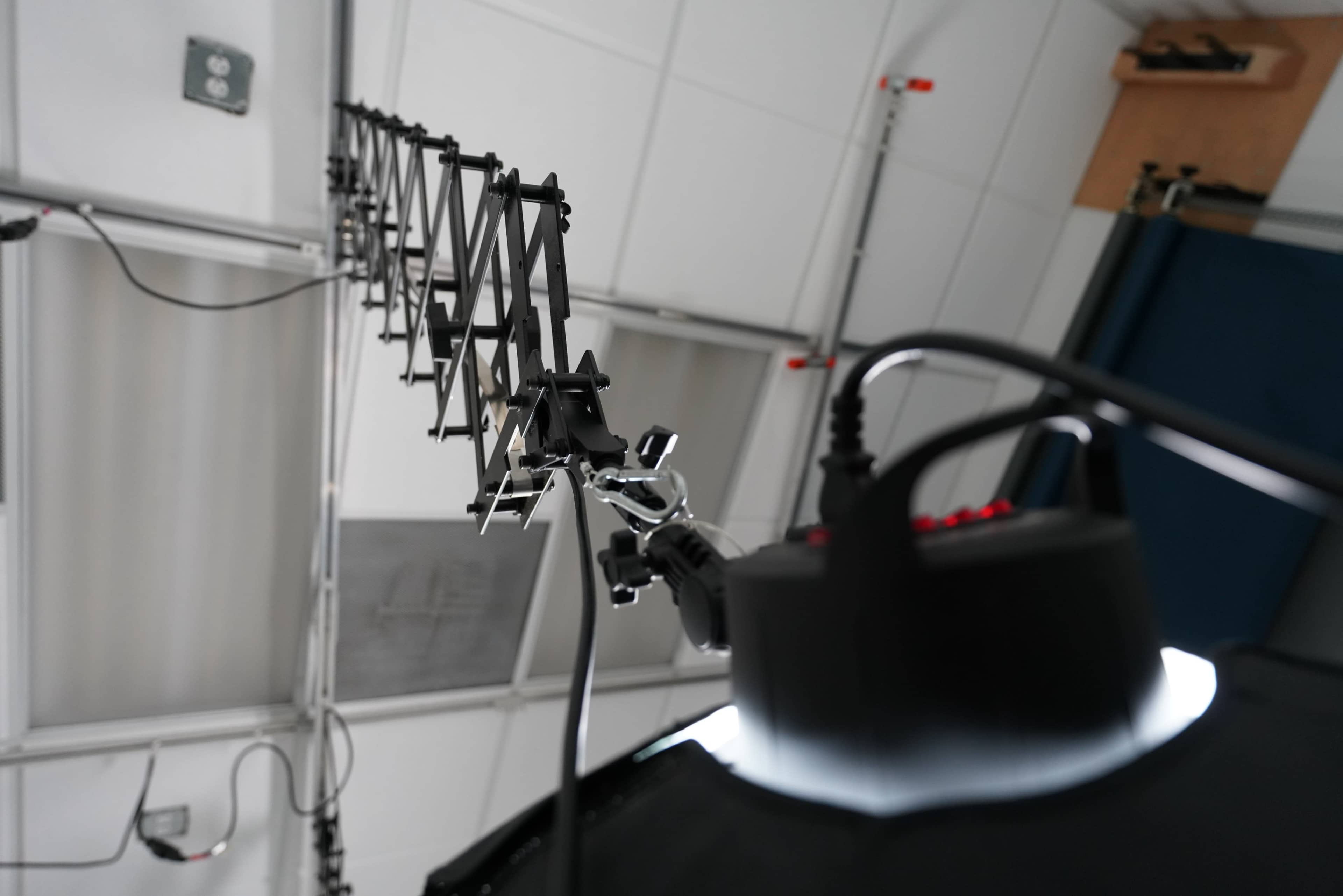 The image shows a close-up view of a black metal structure resembling an antenna, mounted in a laboratory or studio setting, with lights and equipment visible in the background.