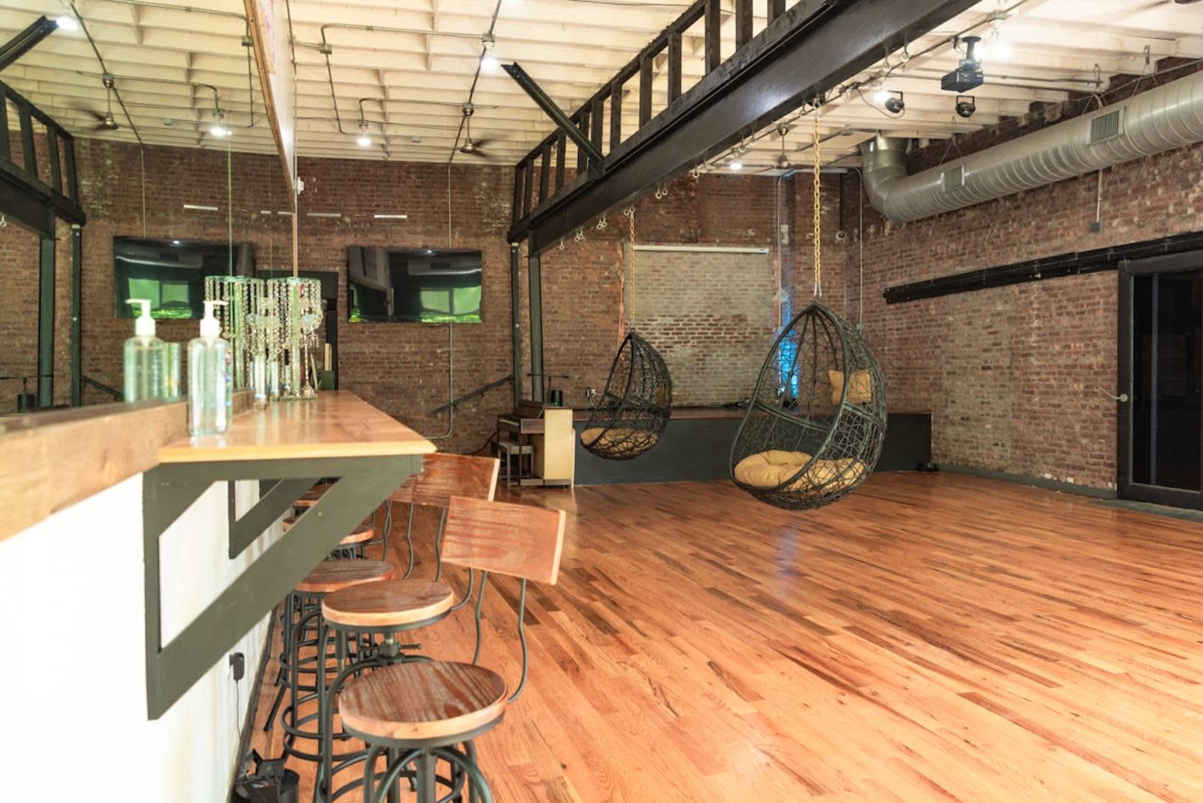 An industrial-style interior with exposed brick walls, a wooden bar with stools, and two hanging chairs.