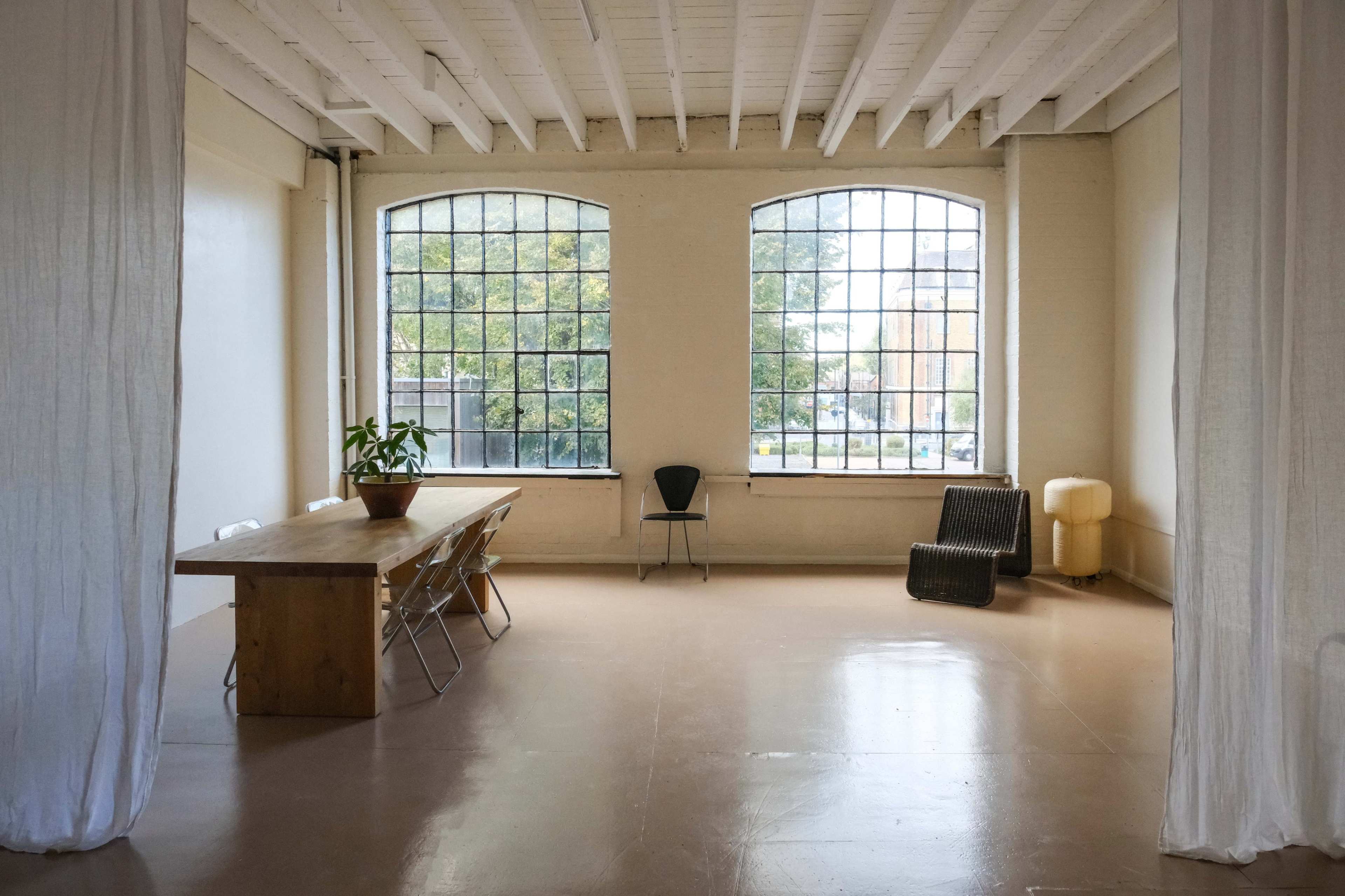 An empty room features a long wooden table, a chair, and large windows that allow natural light to enter.