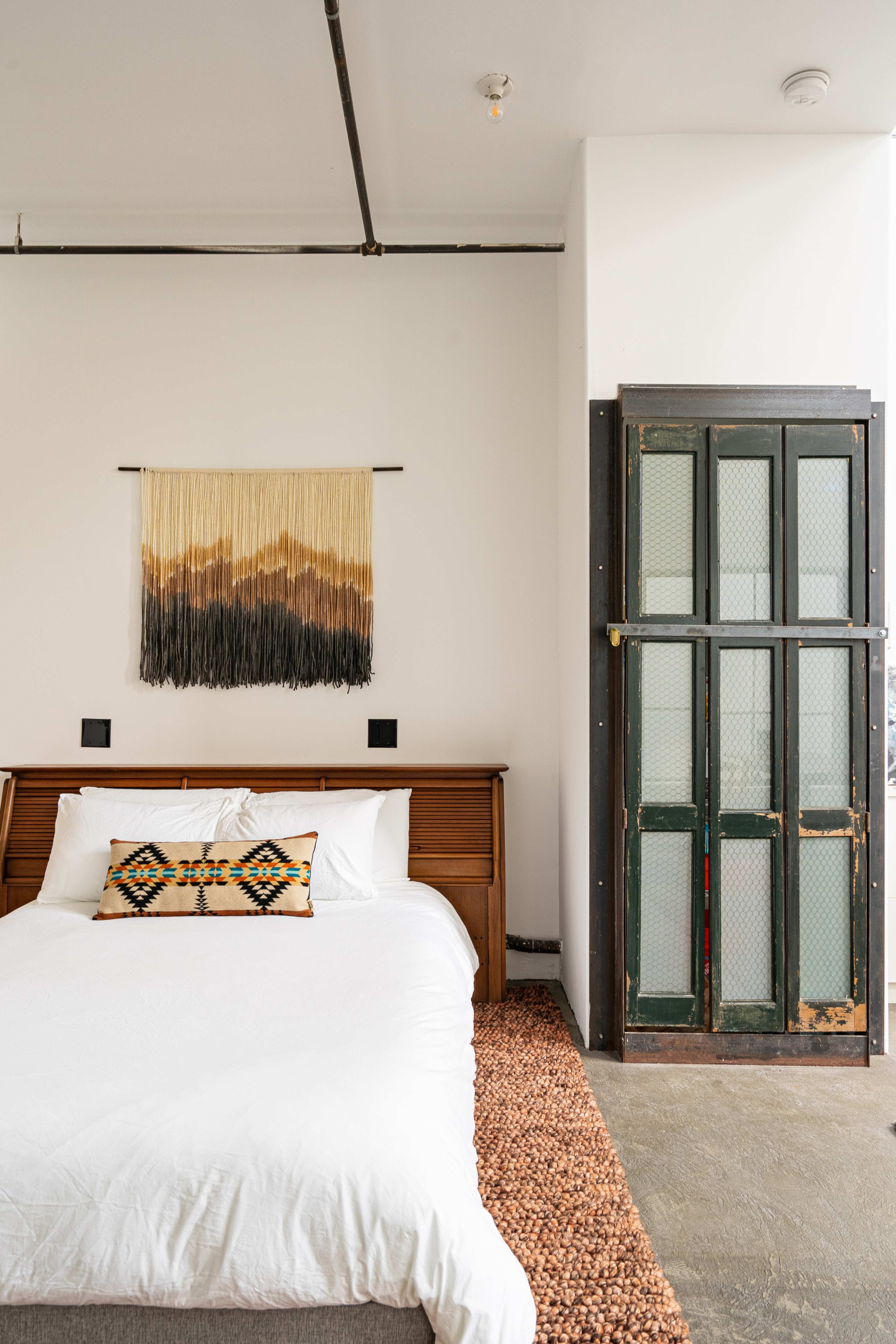 A neatly arranged bedroom features a bed with white bedding, a patterned throw pillow, a woven wall hanging above, and a green door with glass panels.