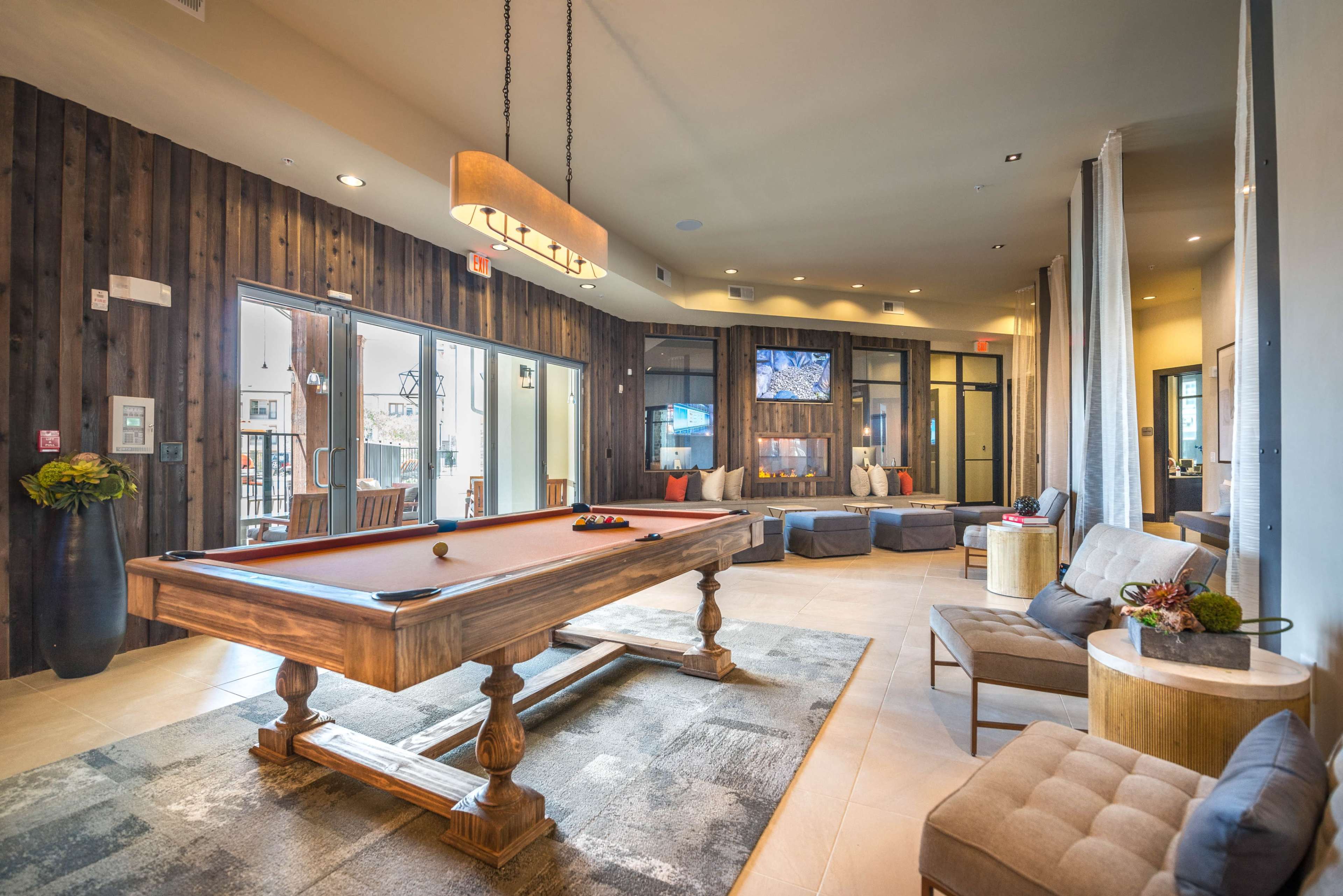 The image shows a recreational lounge area featuring a billiard table, comfortable seating, and large windows that open to an outdoor space.