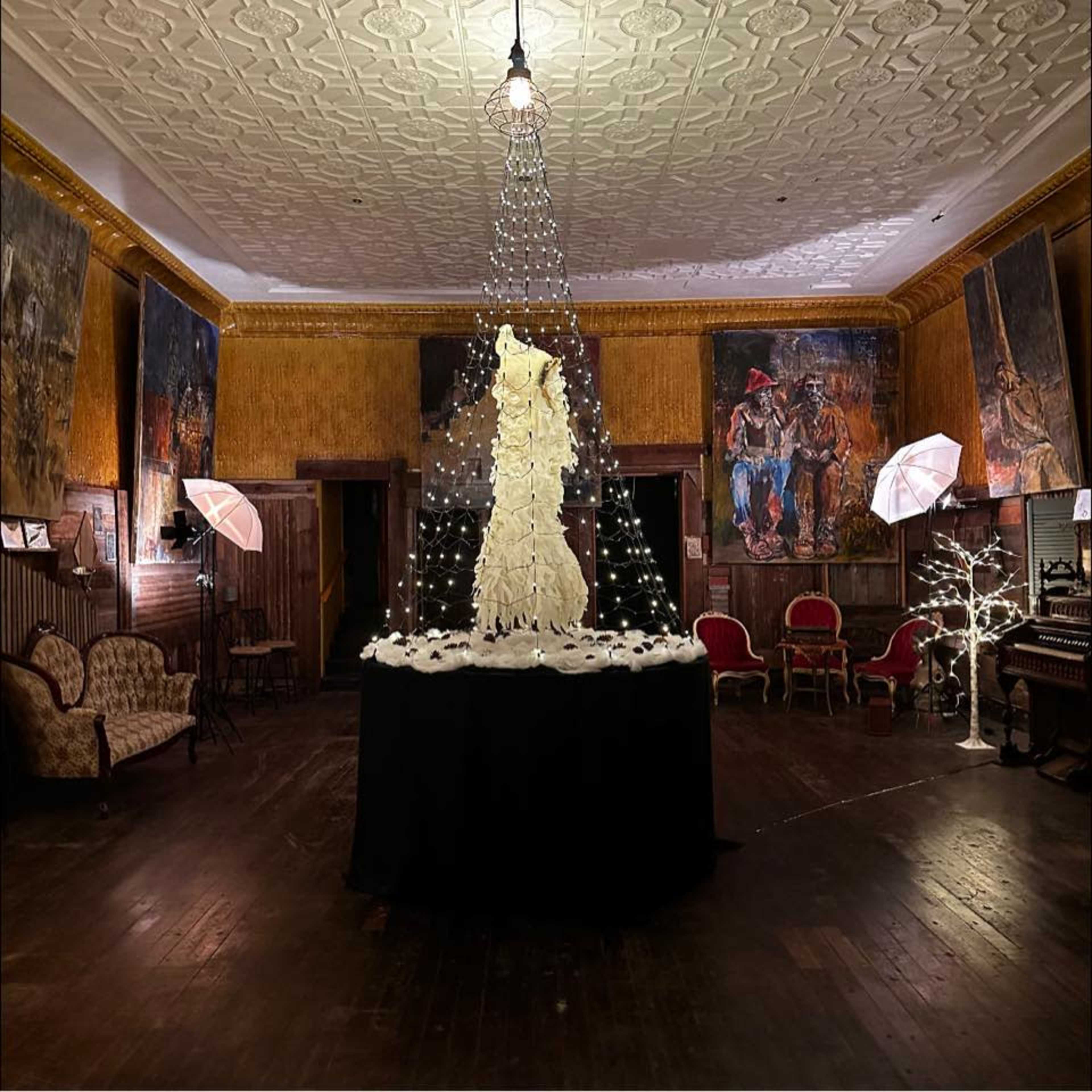 A dimly lit room with a central table draped in black fabric, featuring a tall decorative piece surrounded by lights, and artworks displayed on the walls.