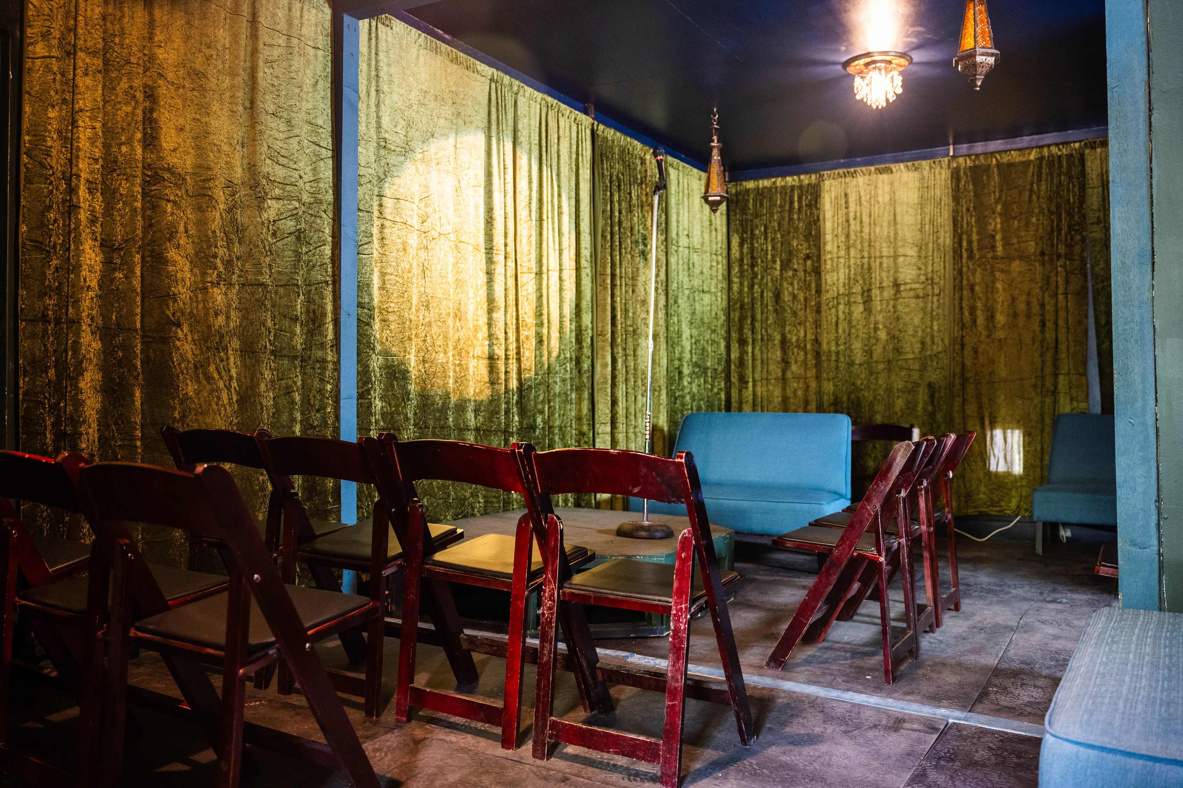 A small, dimly lit room with green curtains, a few blue sofas, and wooden folding chairs arranged around low tables.