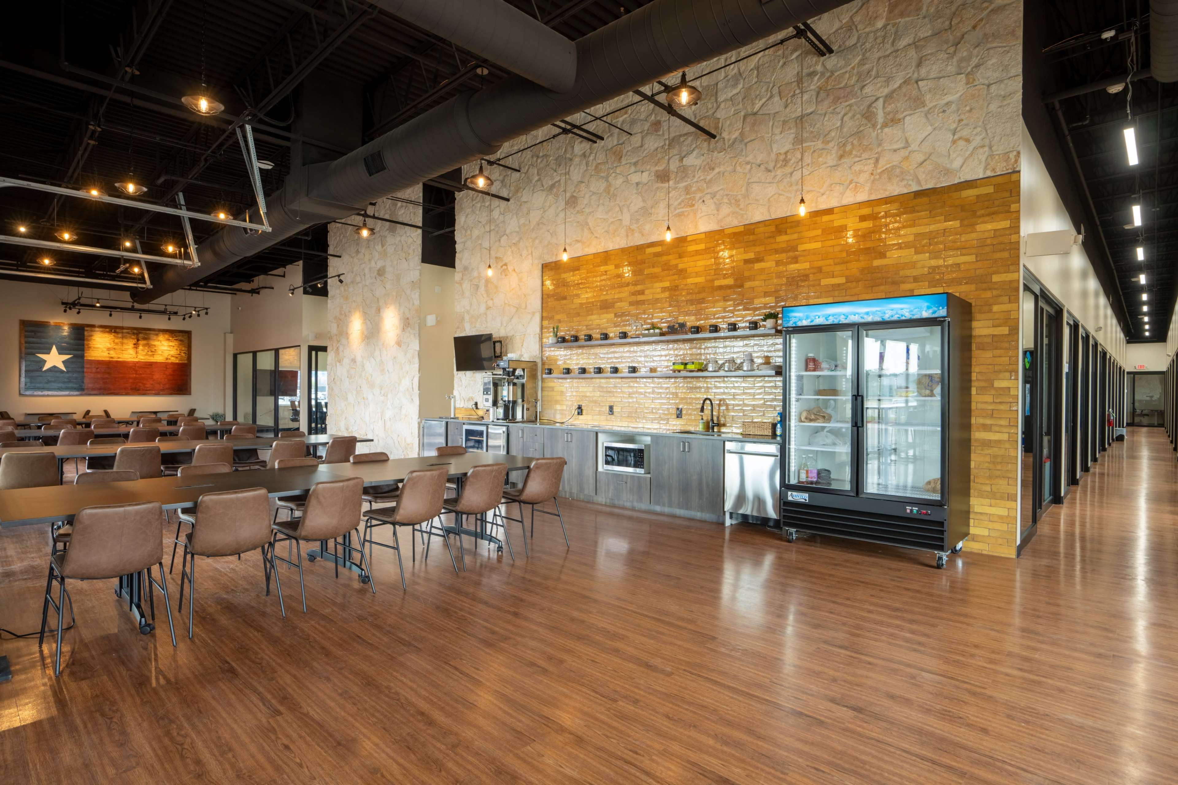 A spacious dining area with rows of tables and chairs, featuring a kitchen counter with a display fridge and a stone wall accent.