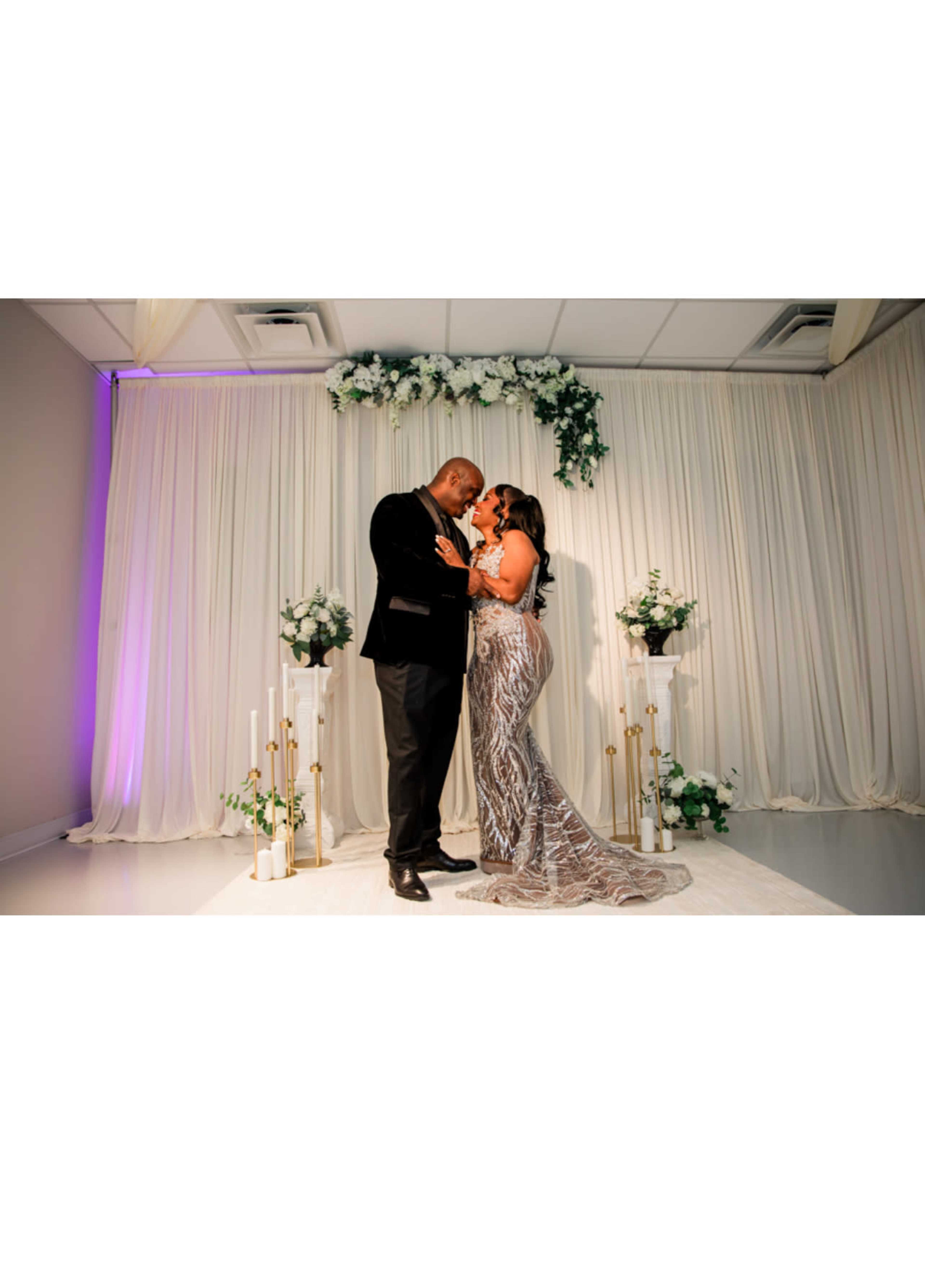 A couple stands in a warmly decorated venue, framed by a decorative backdrop of flowers and candles, sharing an intimate moment.