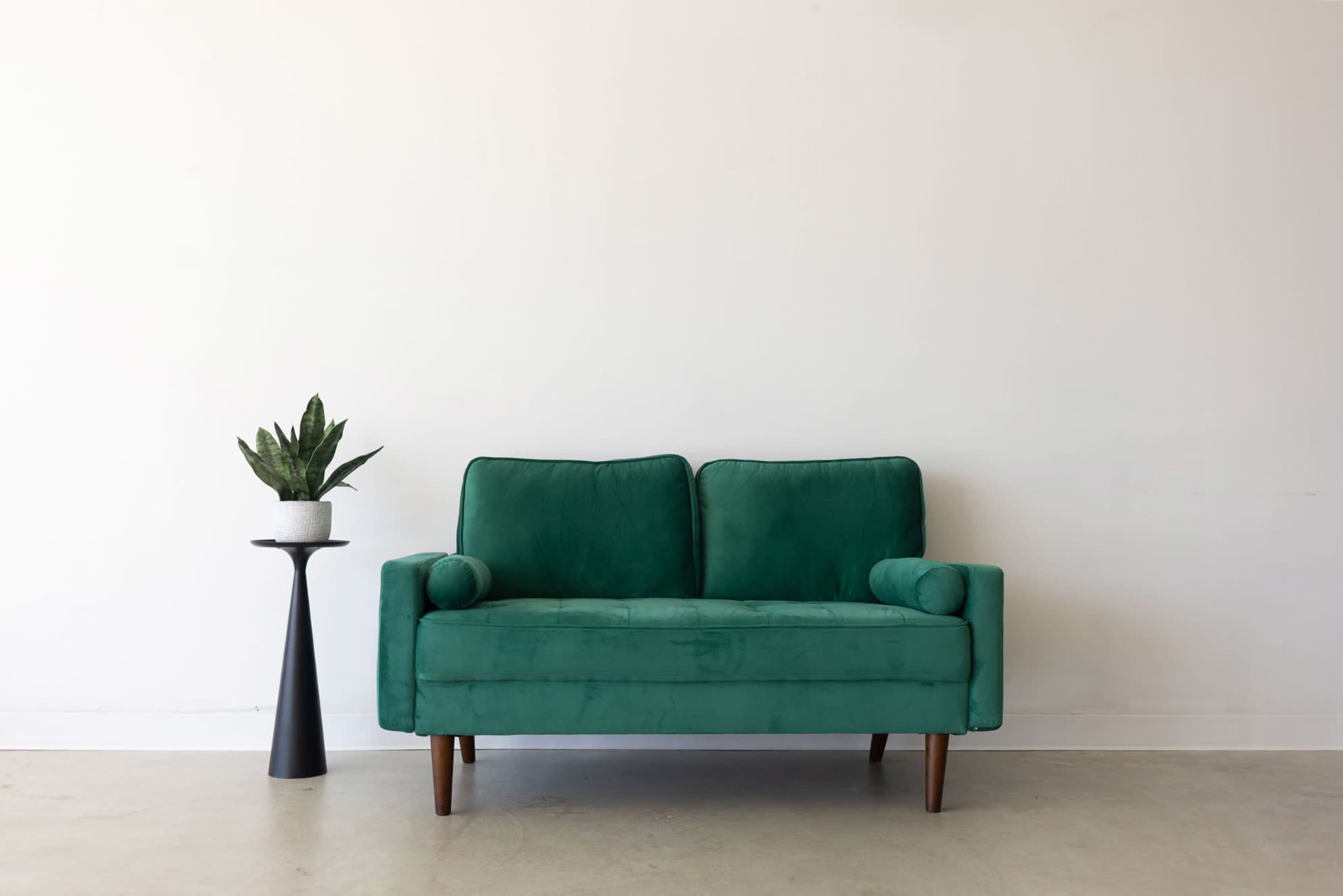 A green velvet loveseat is positioned against a plain white wall, accompanied by a black side table with a potted plant.