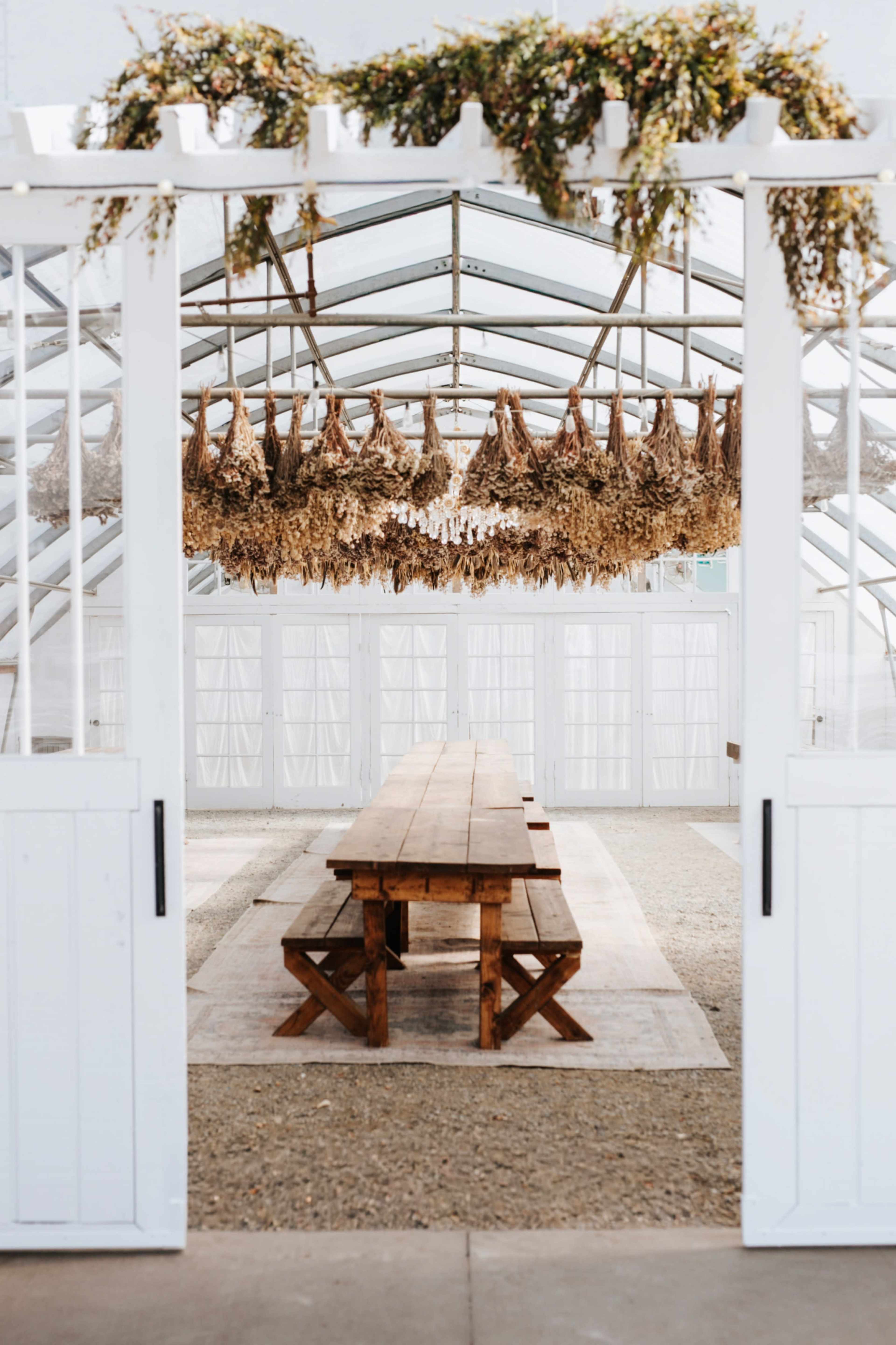 A wooden table with benches is centered in a bright greenhouse, featuring dried floral arrangements hanging overhead.