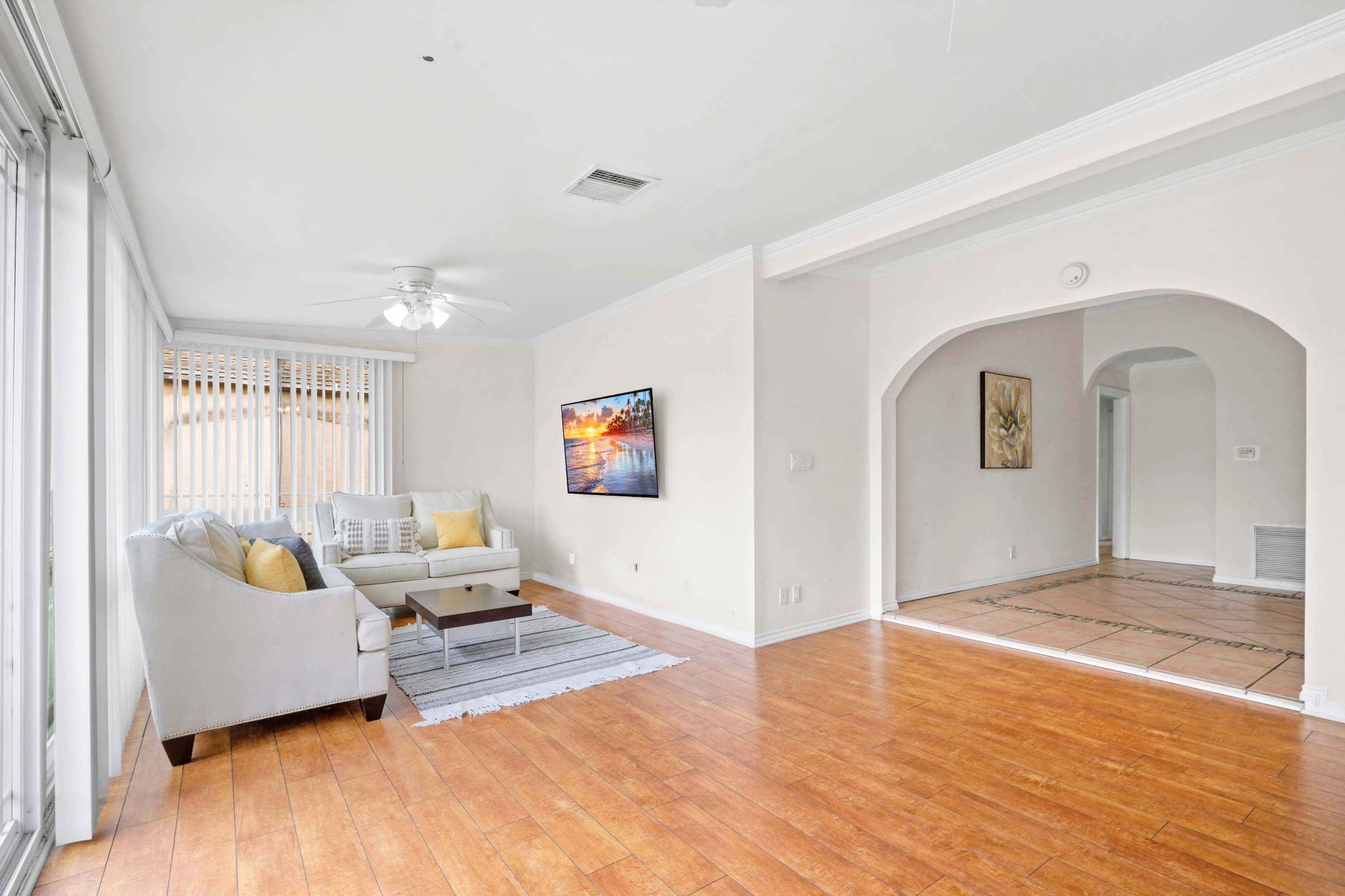 A bright living room features a light-colored sofa, a coffee table, and a wall-mounted television, with an adjoining archway leading to another room.