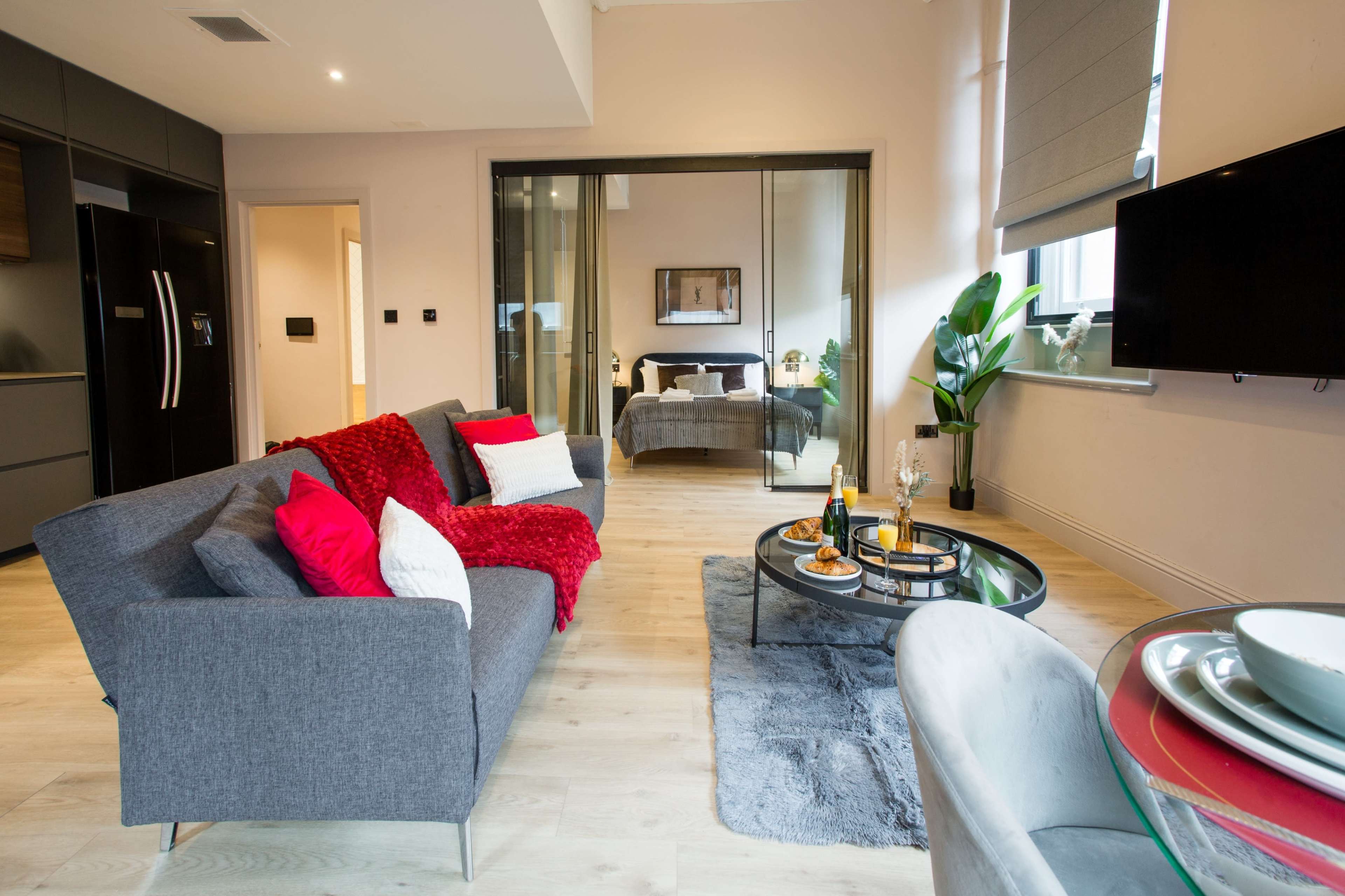 The image shows a modern living room with a gray sofa, a glass coffee table, and a view into a bedroom area through sliding glass doors.