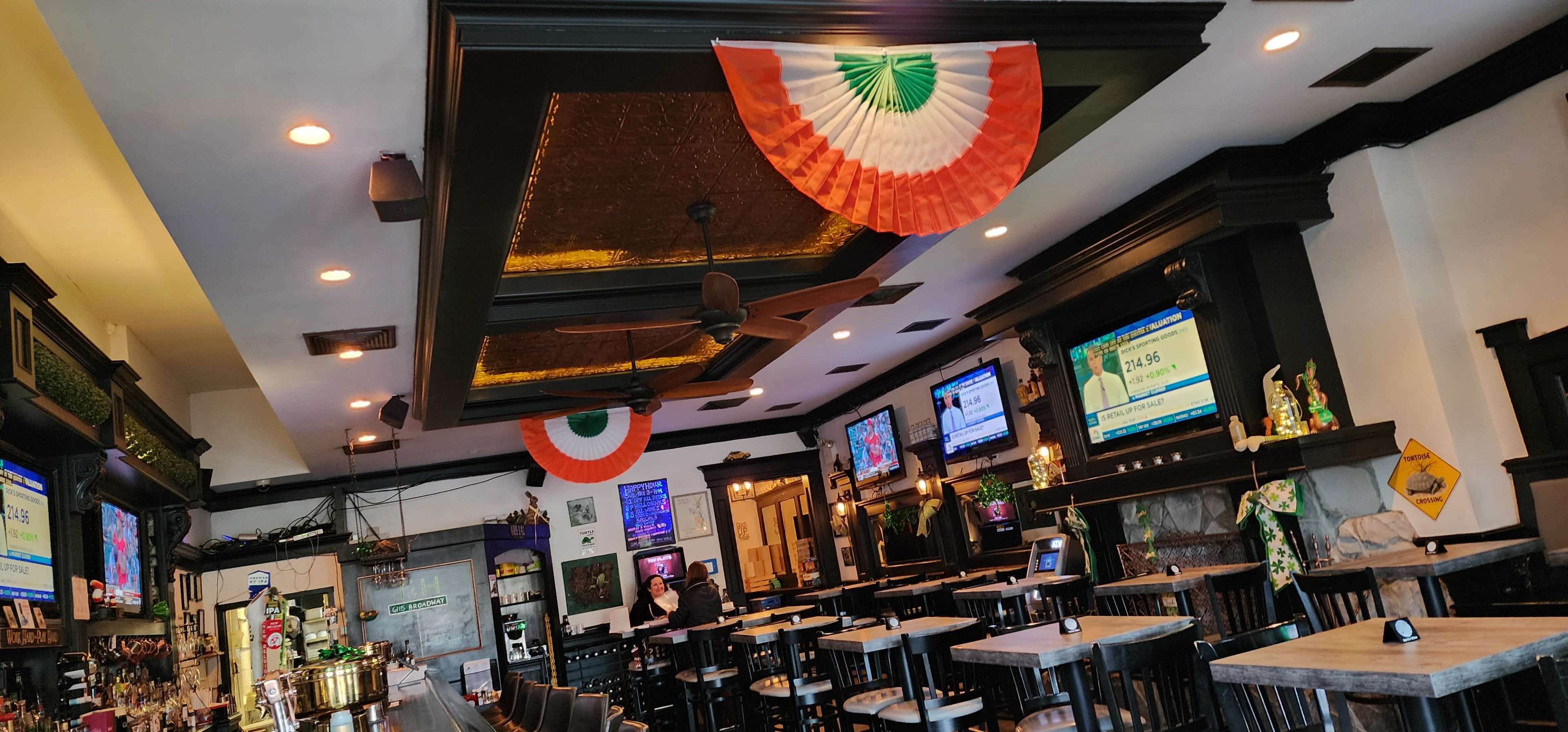 The interior of a bar features multiple television screens, wooden tables and chairs, and decorative Indian flags hanging from the ceiling.