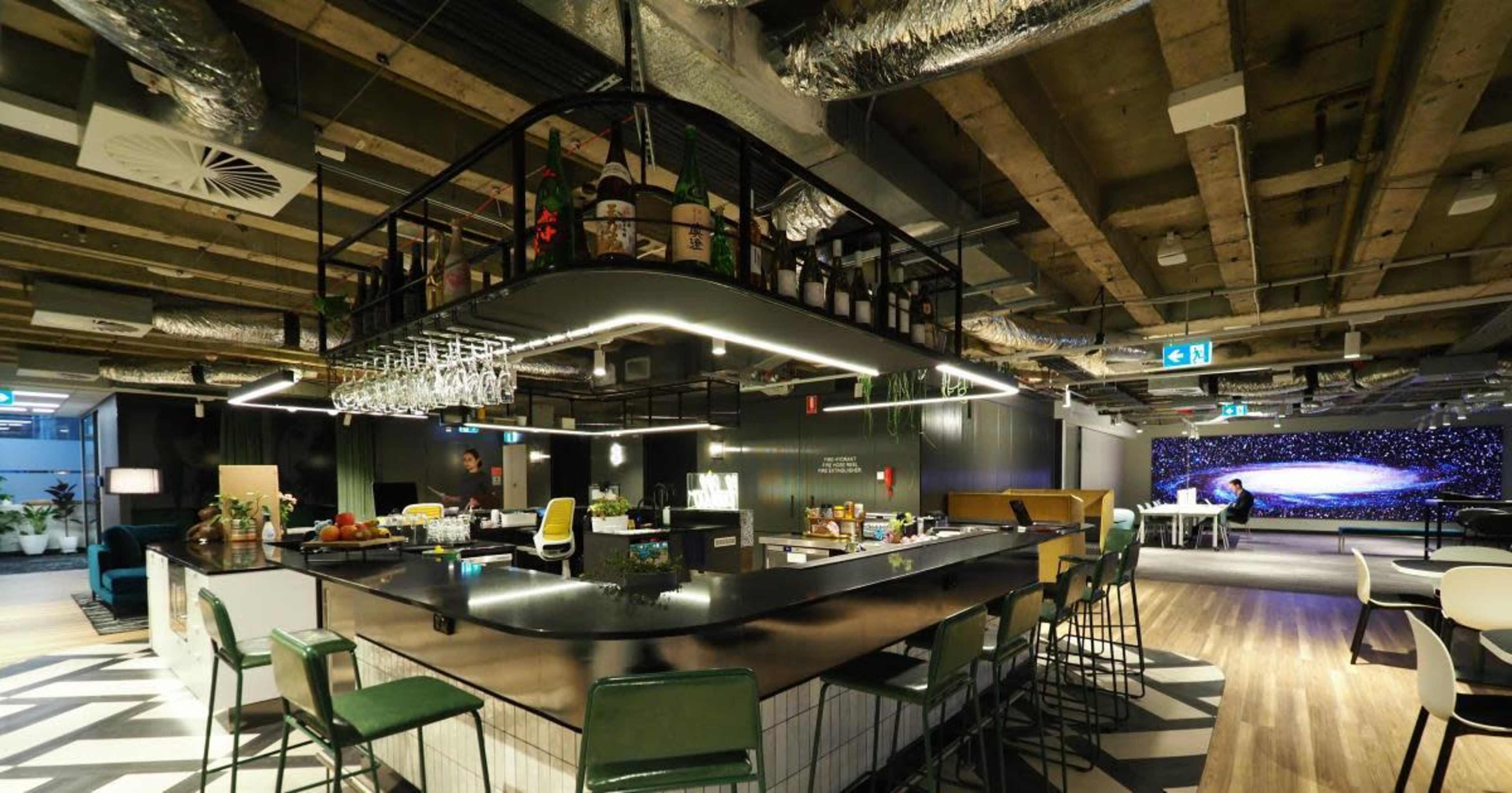 The image shows a modern office space featuring a central bar area with seating, equipped with various beverages and plants, alongside a lounge area and a large artistic wall decoration.