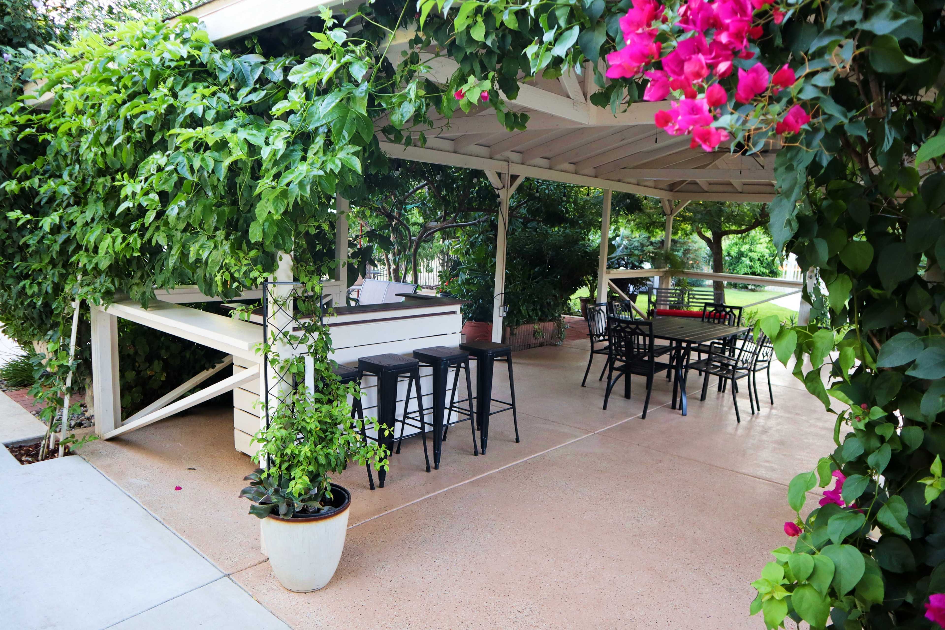 A covered outdoor seating area features black metal chairs and tables surrounded by lush greenery and flowering bougainvillea.