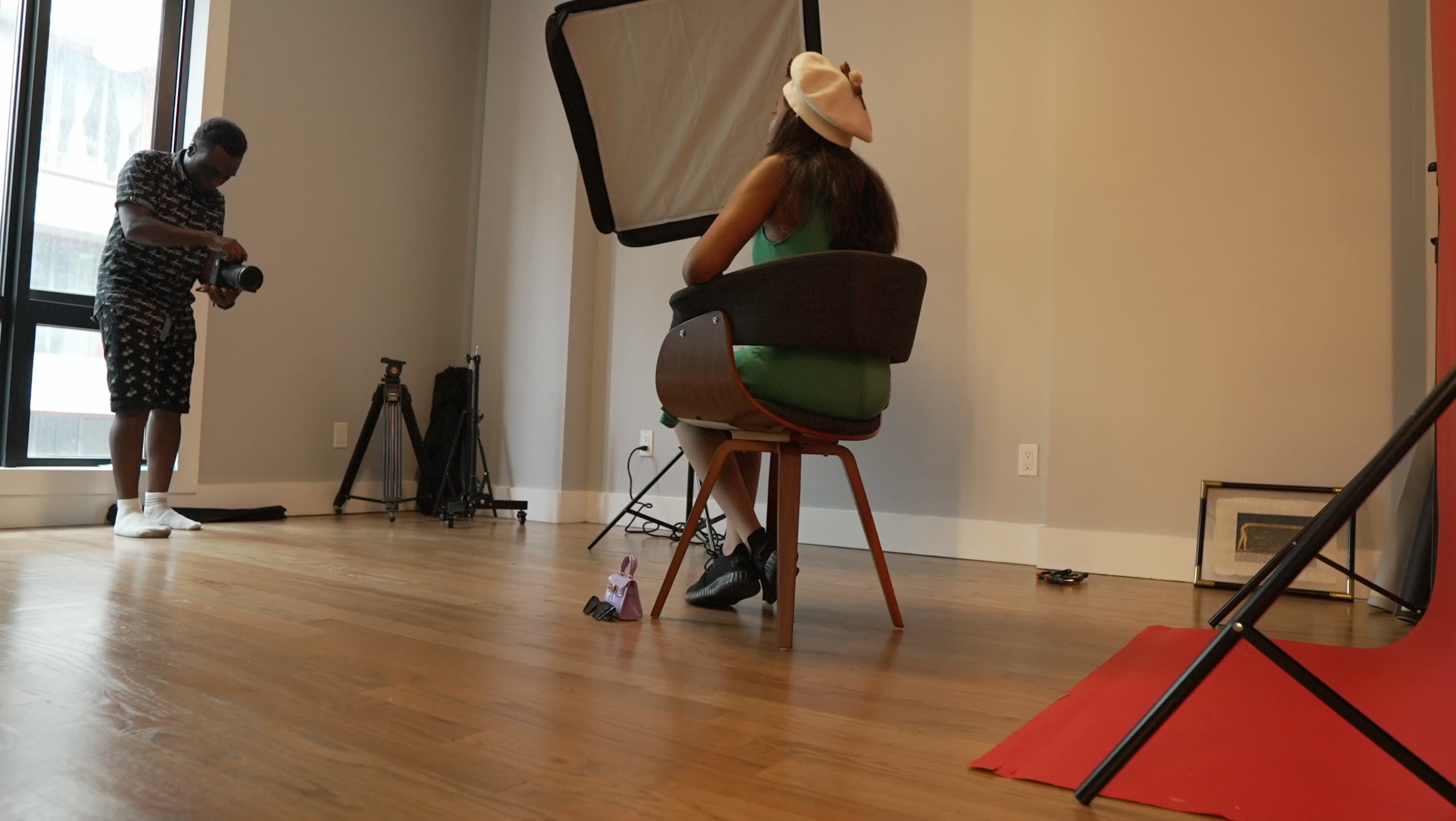 A woman sits in a chair wearing a hat while a man takes her photograph in a well-lit studio.