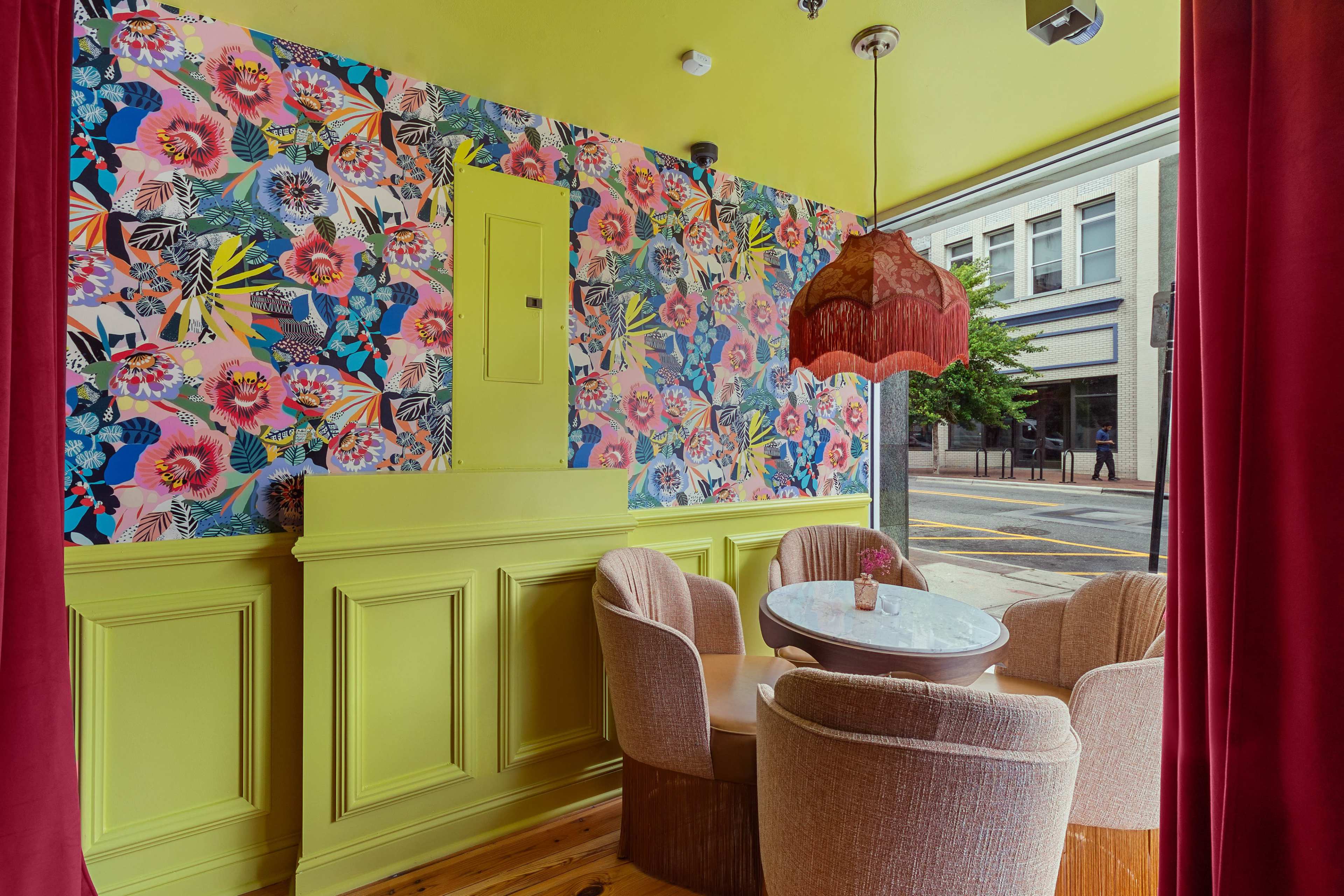The image shows a vibrant cafe interior with floral wallpaper, a lime green wall, and round tables surrounded by plush chairs.