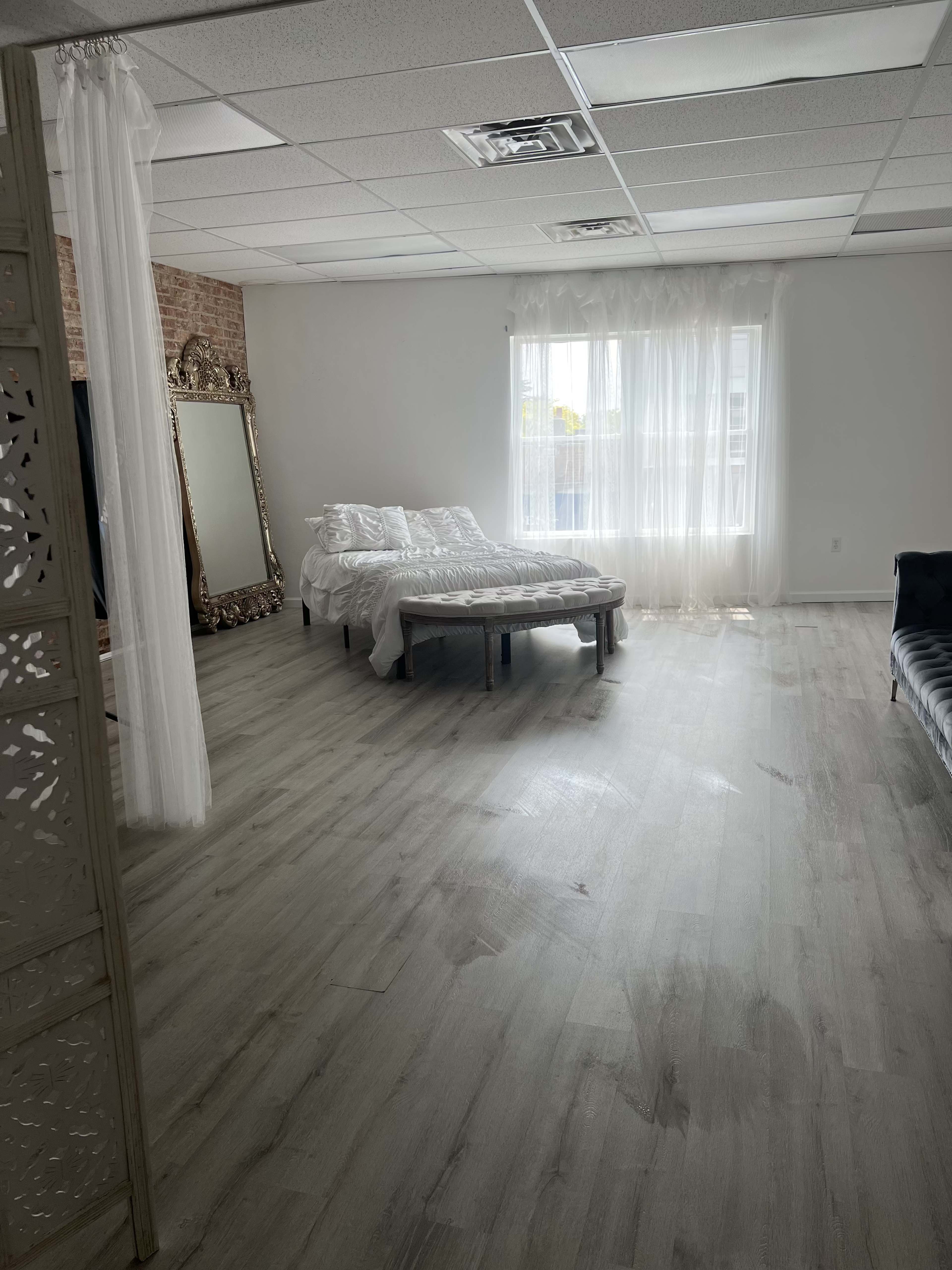 A spacious room with light wooden flooring, a large mirror, a bed covered with white bedding, and sheer curtains allowing natural light to enter.