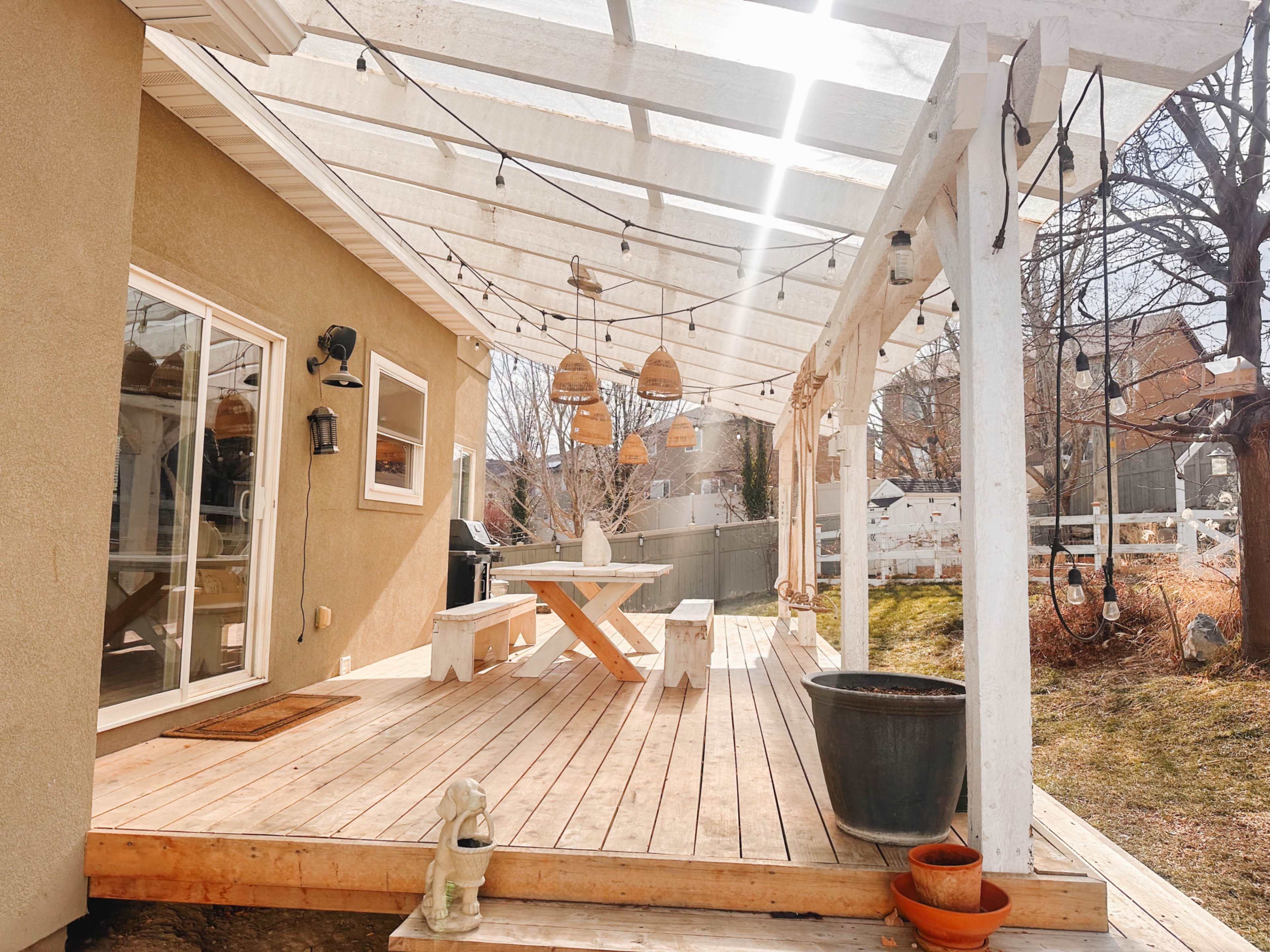 The image shows a wooden patio with a large table and benches, covered by a white pergola, adorned with hanging lights and surrounded by grass and trees.