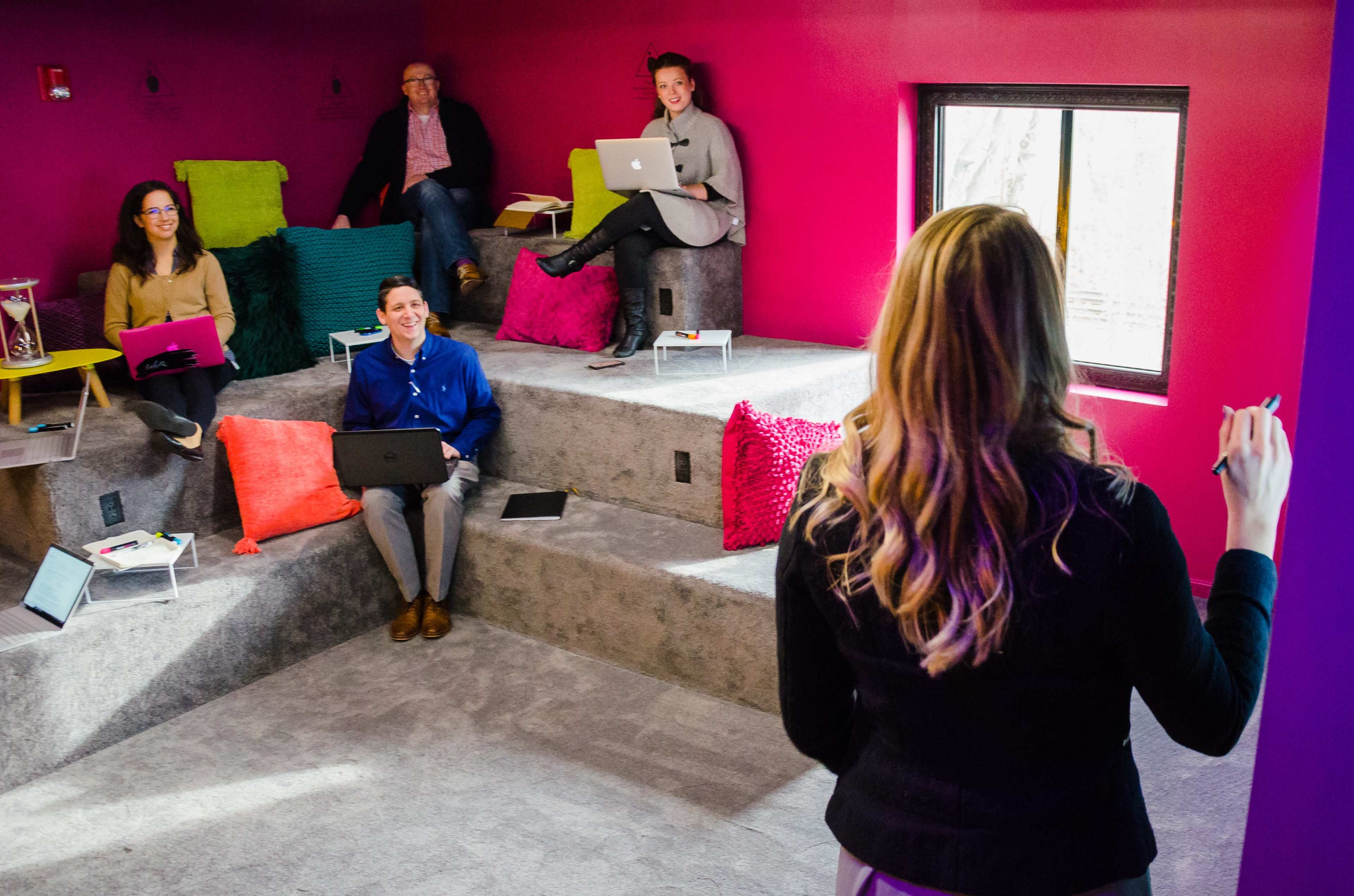 A group of five people are seated in a colorful, tiered seating area, engaged in a meeting while using laptops.