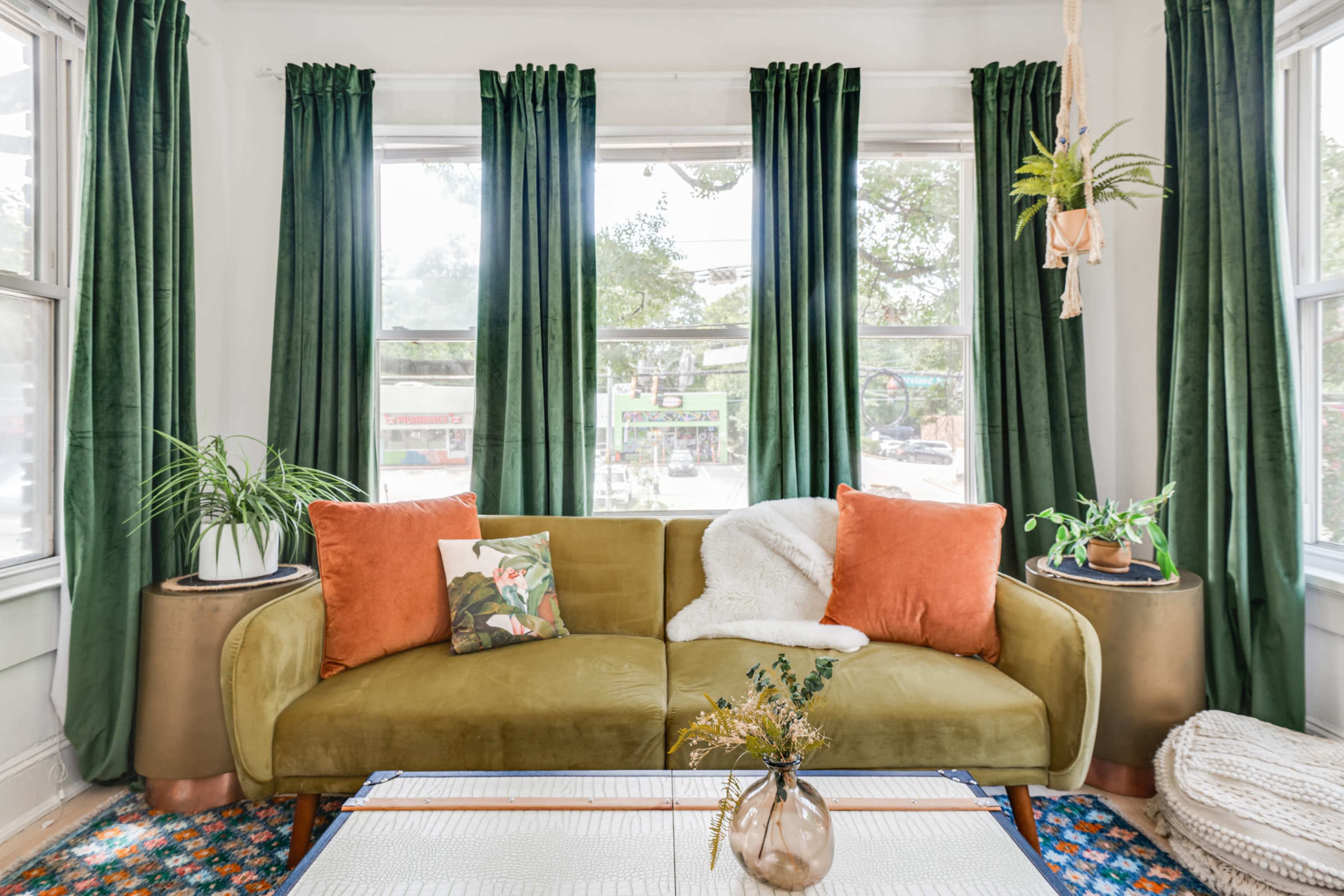 The image shows a stylish living room featuring a green velvet sofa with orange cushions, two potted plants, and large windows adorned with long green curtains.