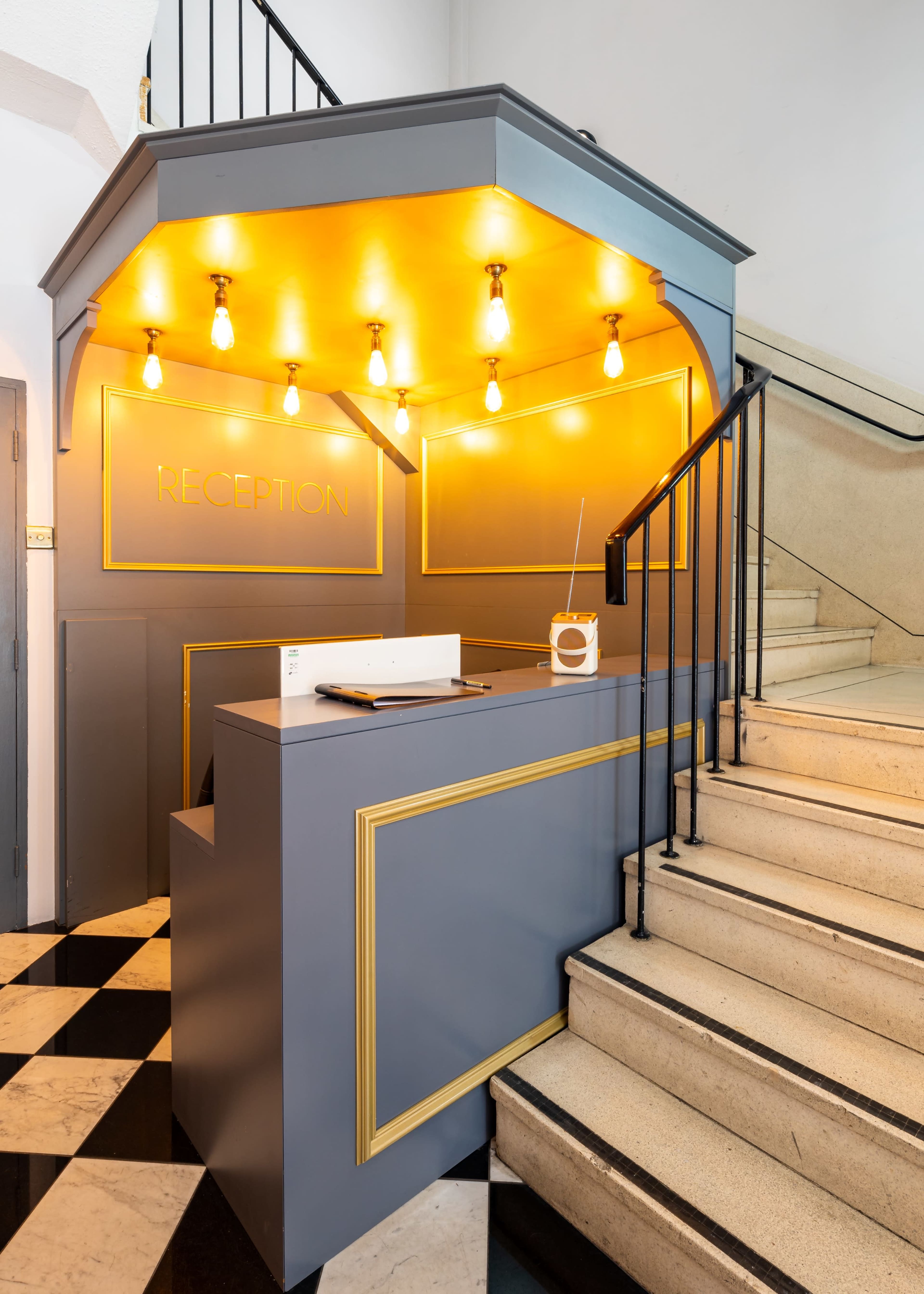 A reception desk with a gray and gold design, illuminated by exposed light bulbs, located next to a staircase with black and white tiled flooring.