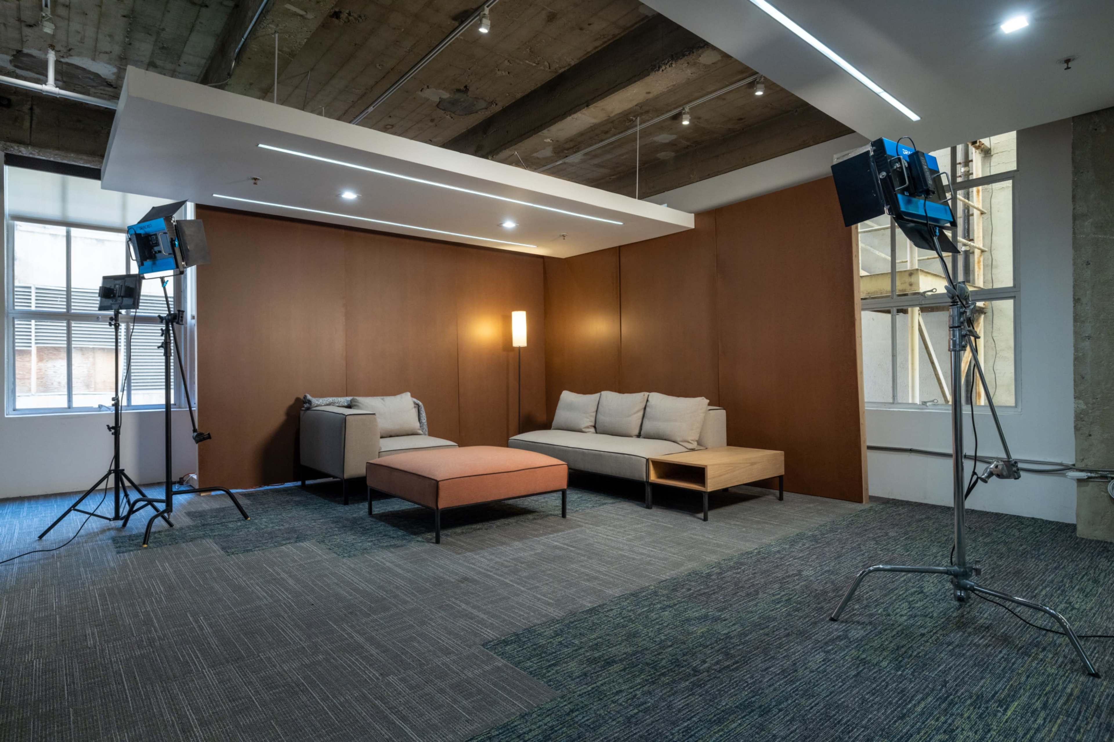 The image shows a modern interior space with a sofa, an ottoman, and two lighting equipment setups in a room with exposed beams and large windows.