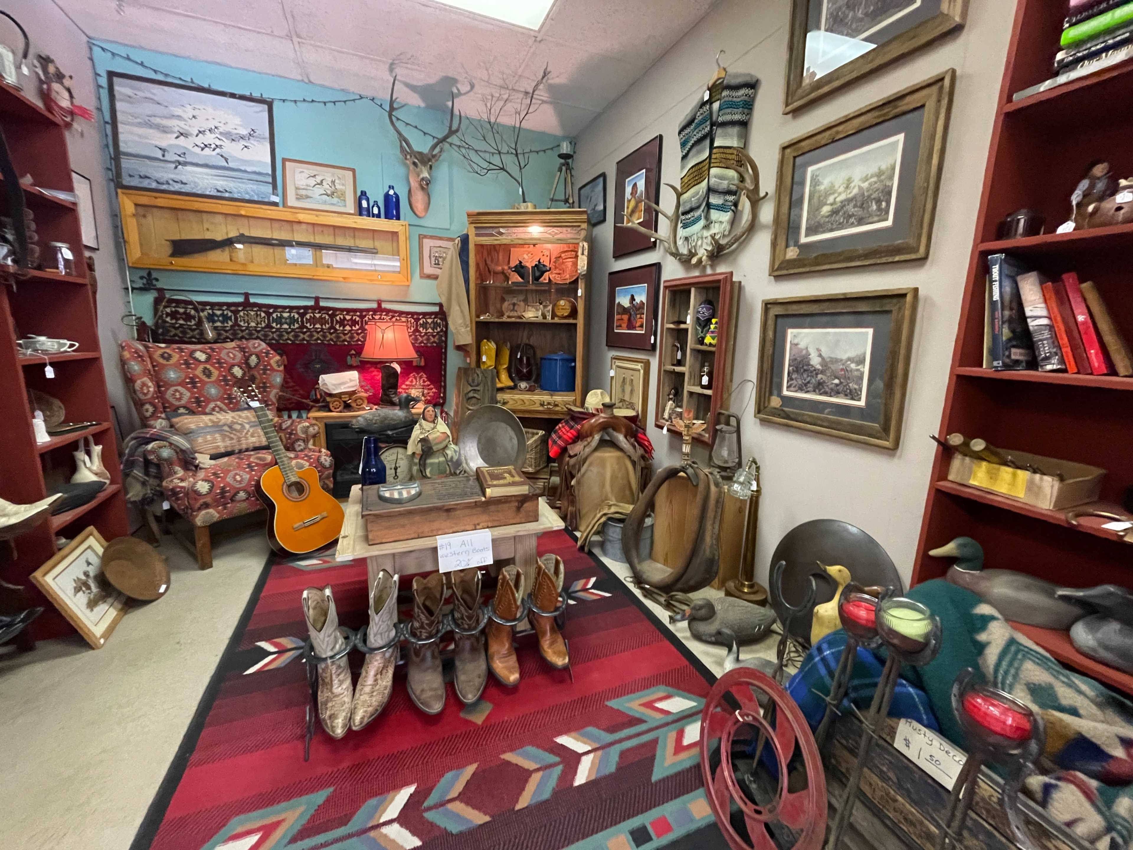 The image shows a cozy antique shop filled with vintage furniture, decorative items, and various collectibles, including a pair of cowboy boots and a guitar.