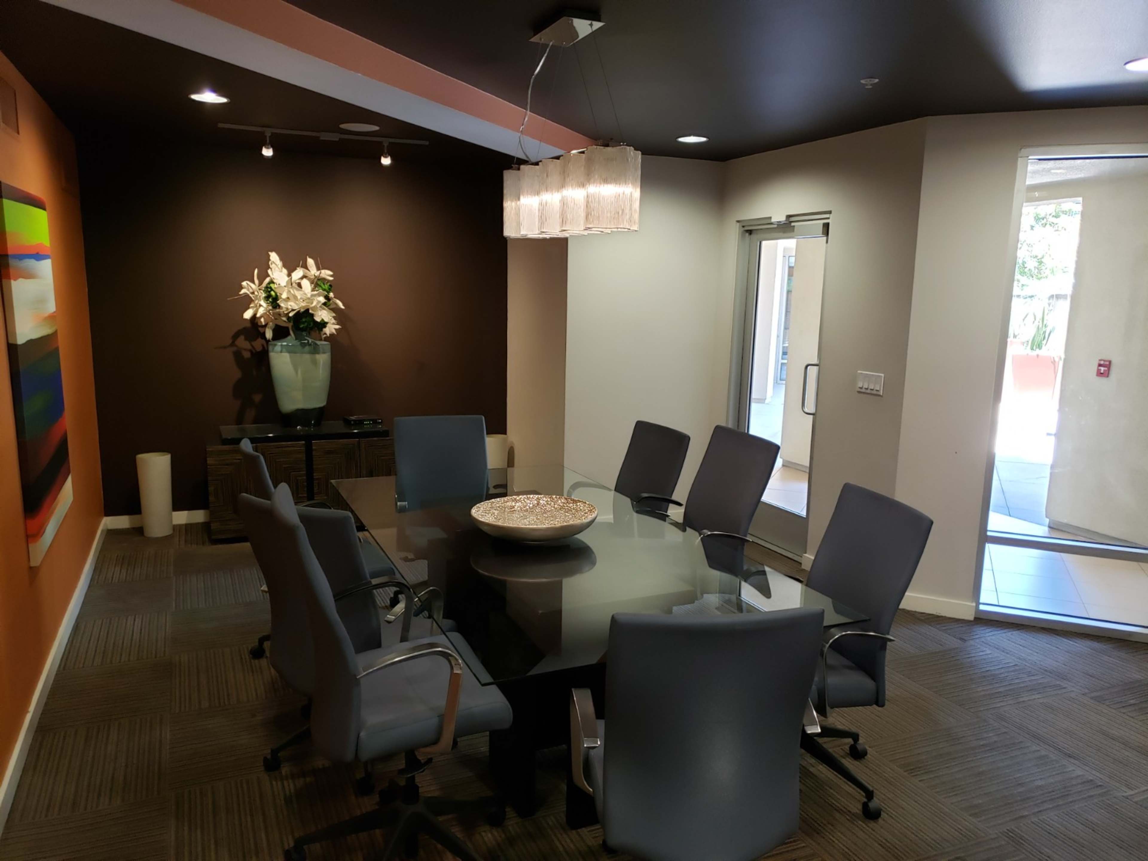 The image shows a modern conference room with a glass table surrounded by eight chairs, a large vase of flowers on the table, and a sliding glass door leading outside.