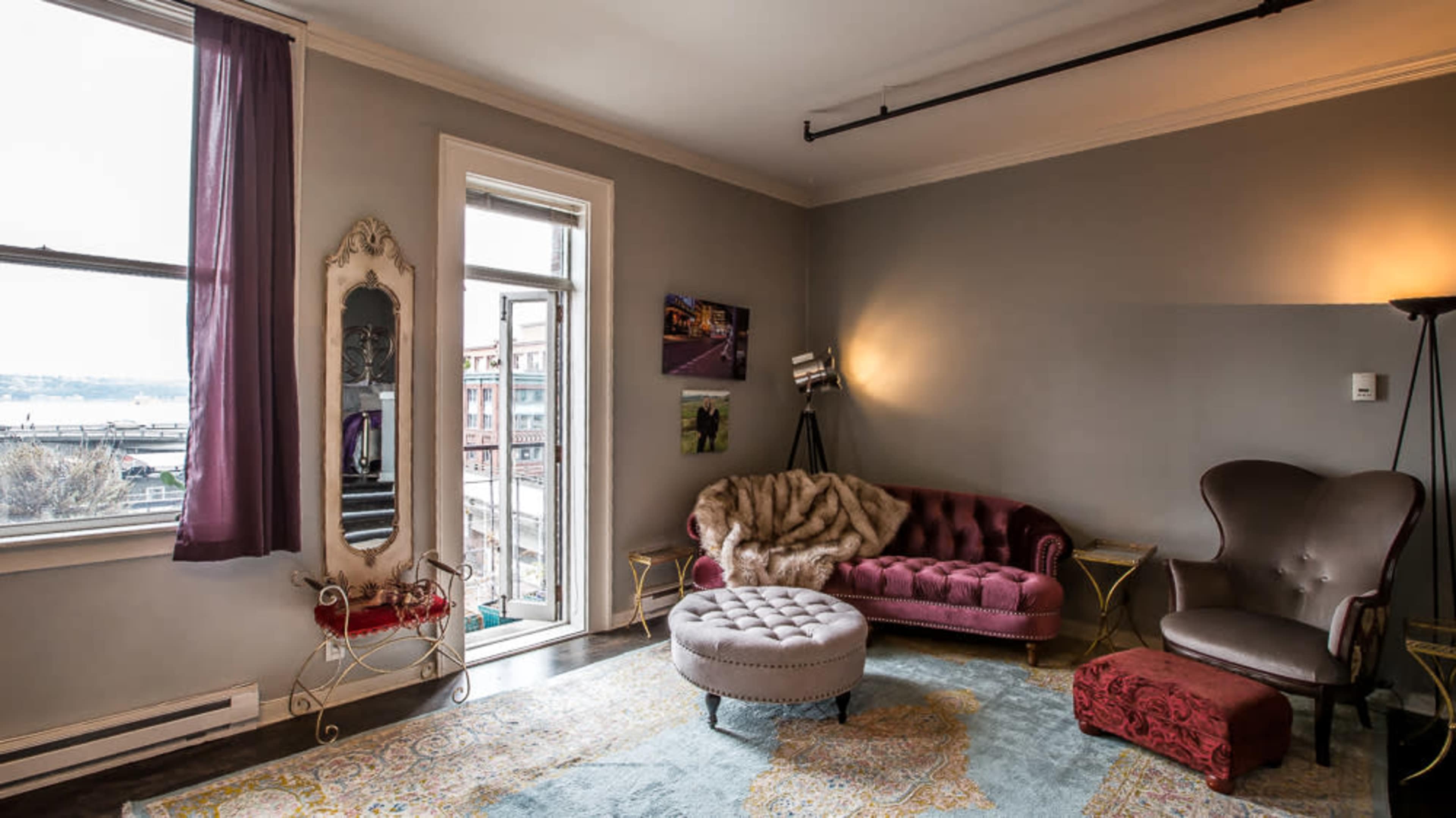 The image shows a cozy living room with a velvet sofa, an ornate mirror, a plush ottoman, and large windows overlooking a cityscape.