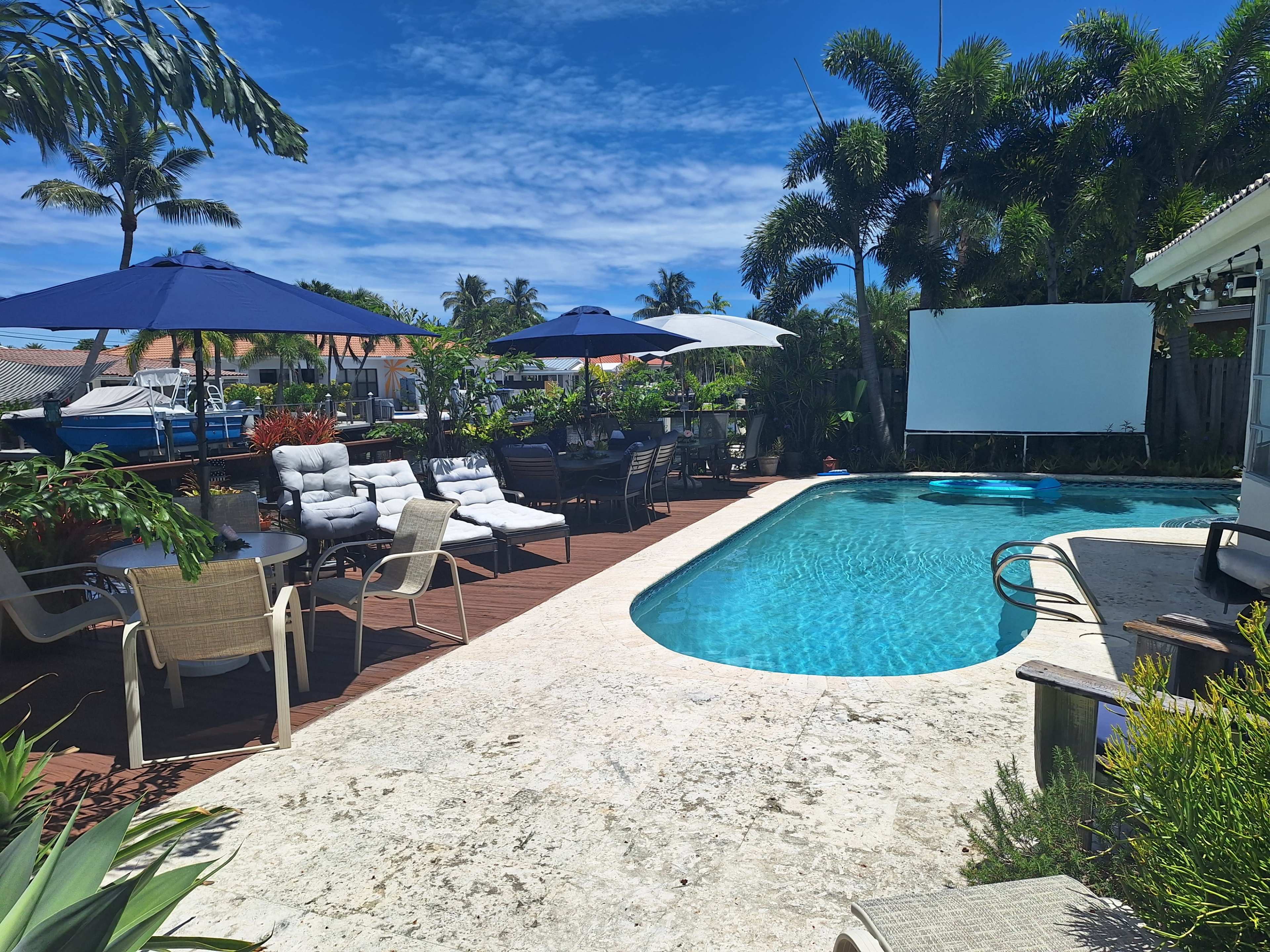The scene features a backyard pool area with sun loungers and umbrellas, surrounded by tropical vegetation and a large screen positioned near the pool.