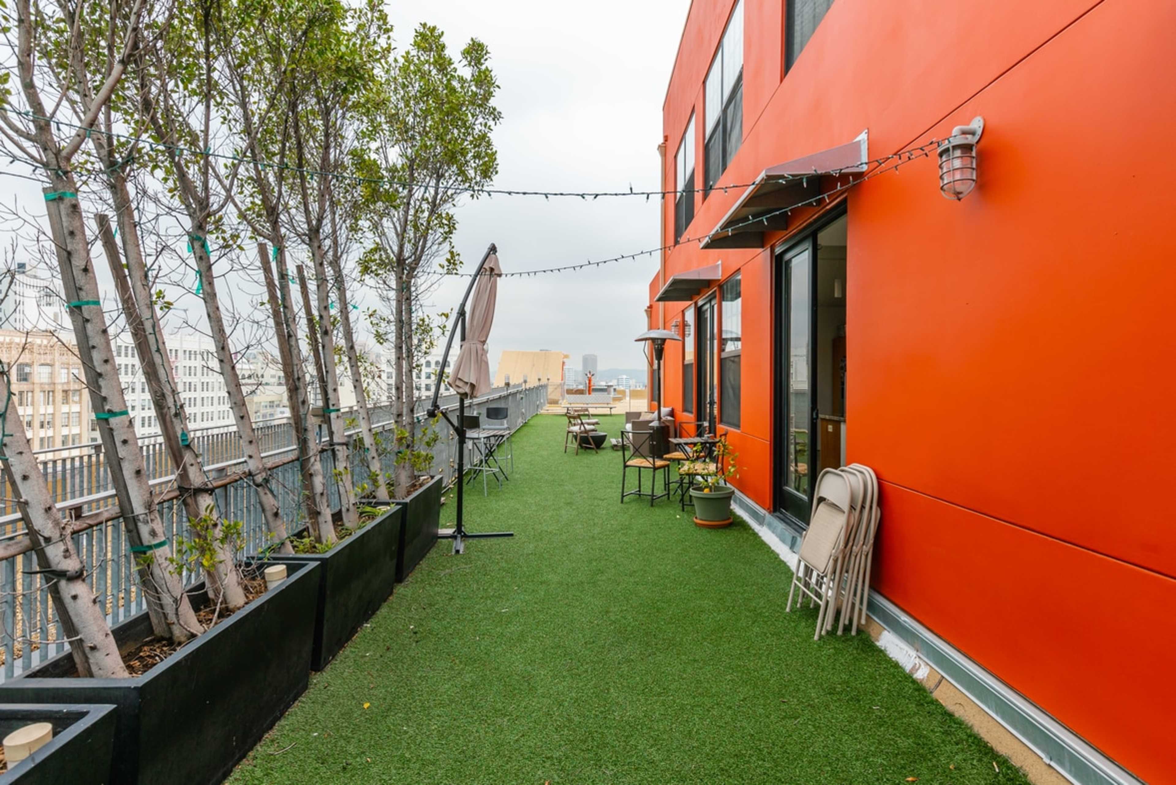 A rooftop terrace with artificial grass, potted plants, and folding chairs along the orange-colored building wall.