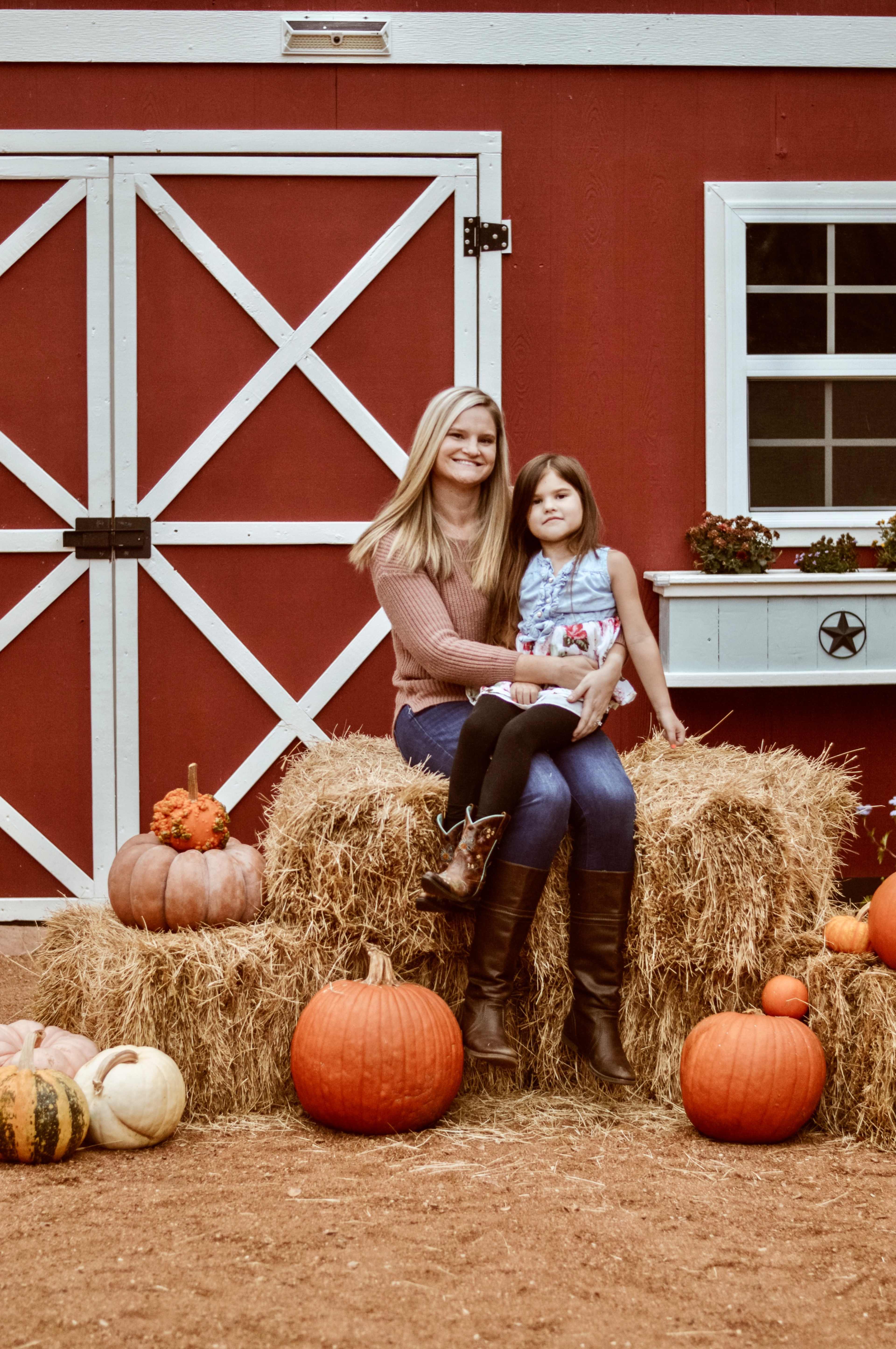 A woman and a child sit on a stack of hay bales in front of a red barn, surrounded by pumpkins and gourds.