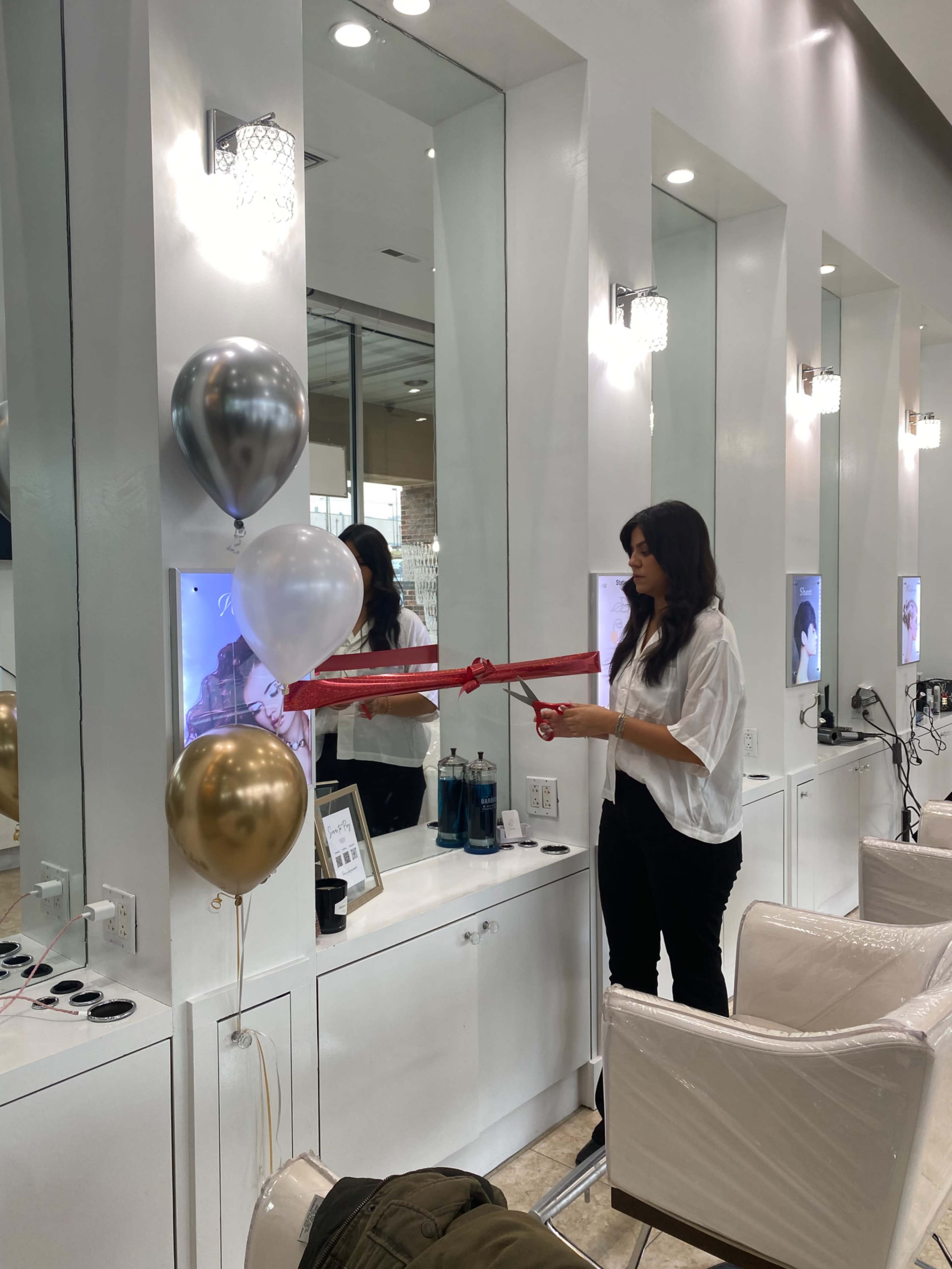 A woman dressed in a white shirt ties a ribbon around a display in a hair salon that features balloons and beauty products.