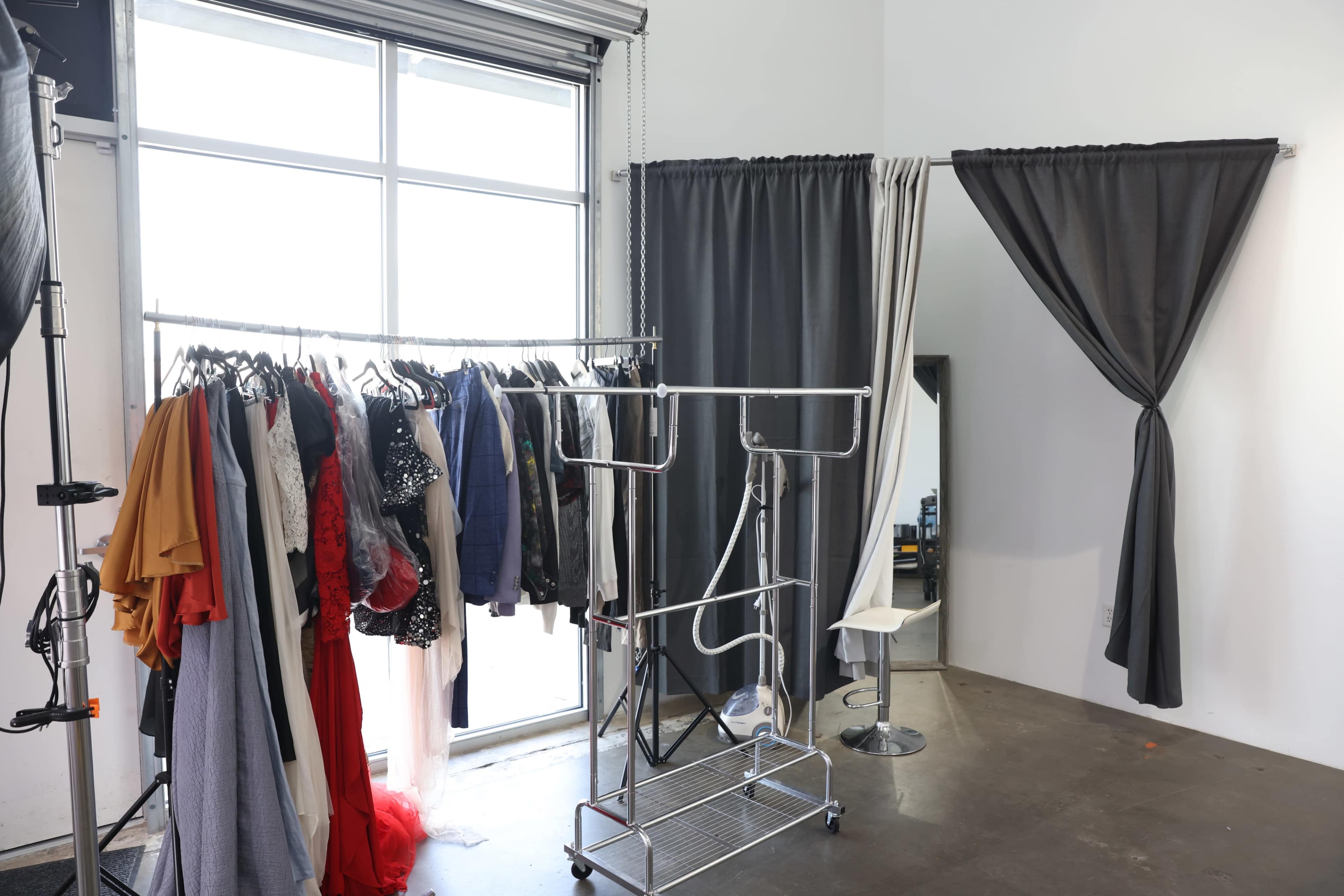 A clothing rack filled with various garments next to a large window and a mirror surrounded by curtains.